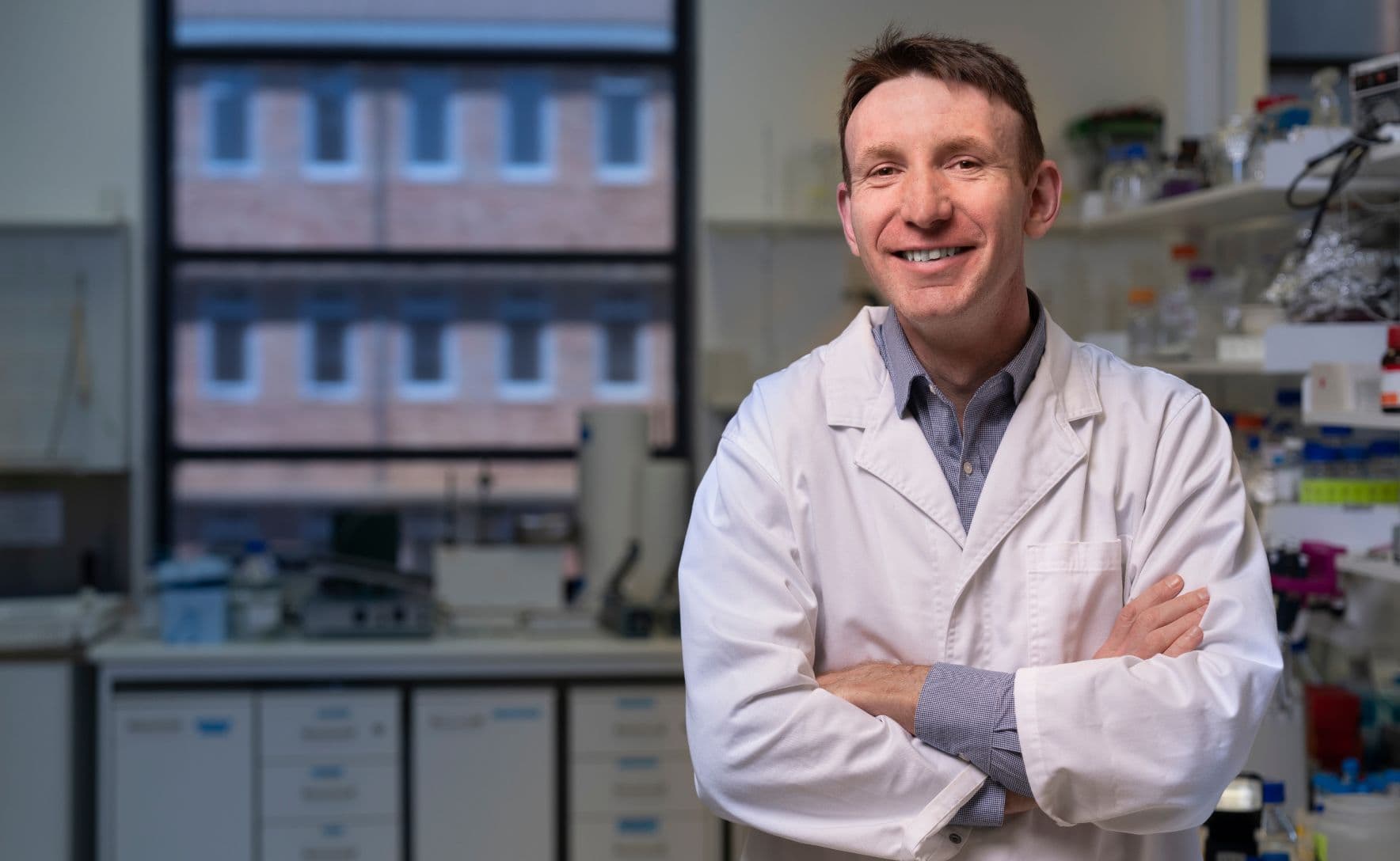 Ian is wearing a white lab coat and smiling at the camera with his arms crossed. e is standing in a laboratory with various scientific equipment in the background