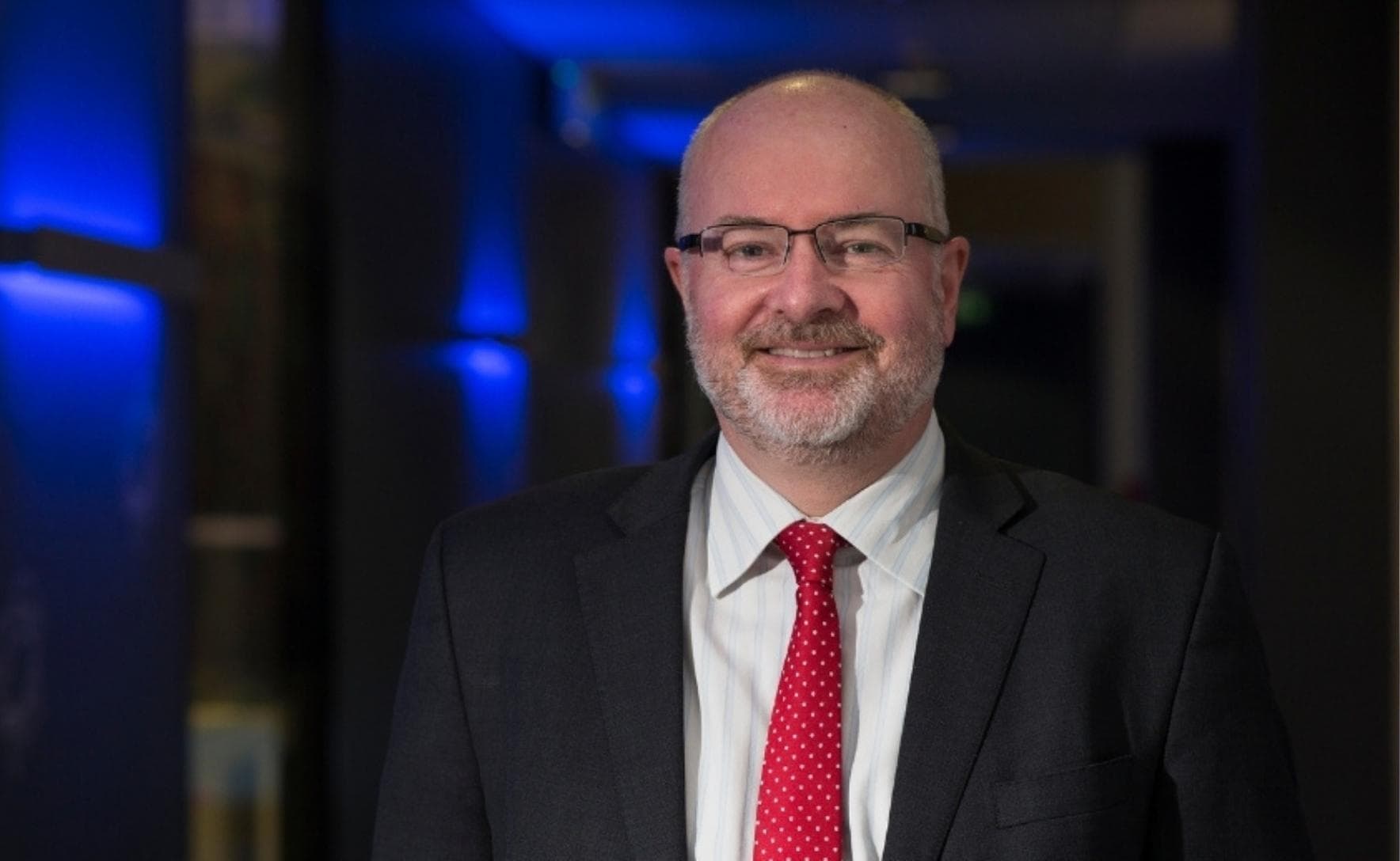 Ross is is standing in front of a dark corridor lit by blue lights. He is wearing a black suit, white collared shirt and red tie. He is smiling at the camera. 