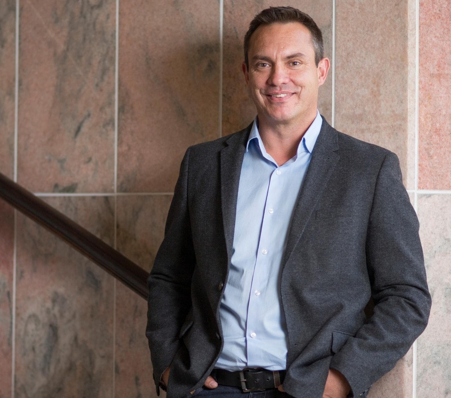 Ray smiles at the camera and wears a dark grey suit jacket and light blue collared shirt. He is standing on a staircase. Behind him is a wall of brown stone tiles.