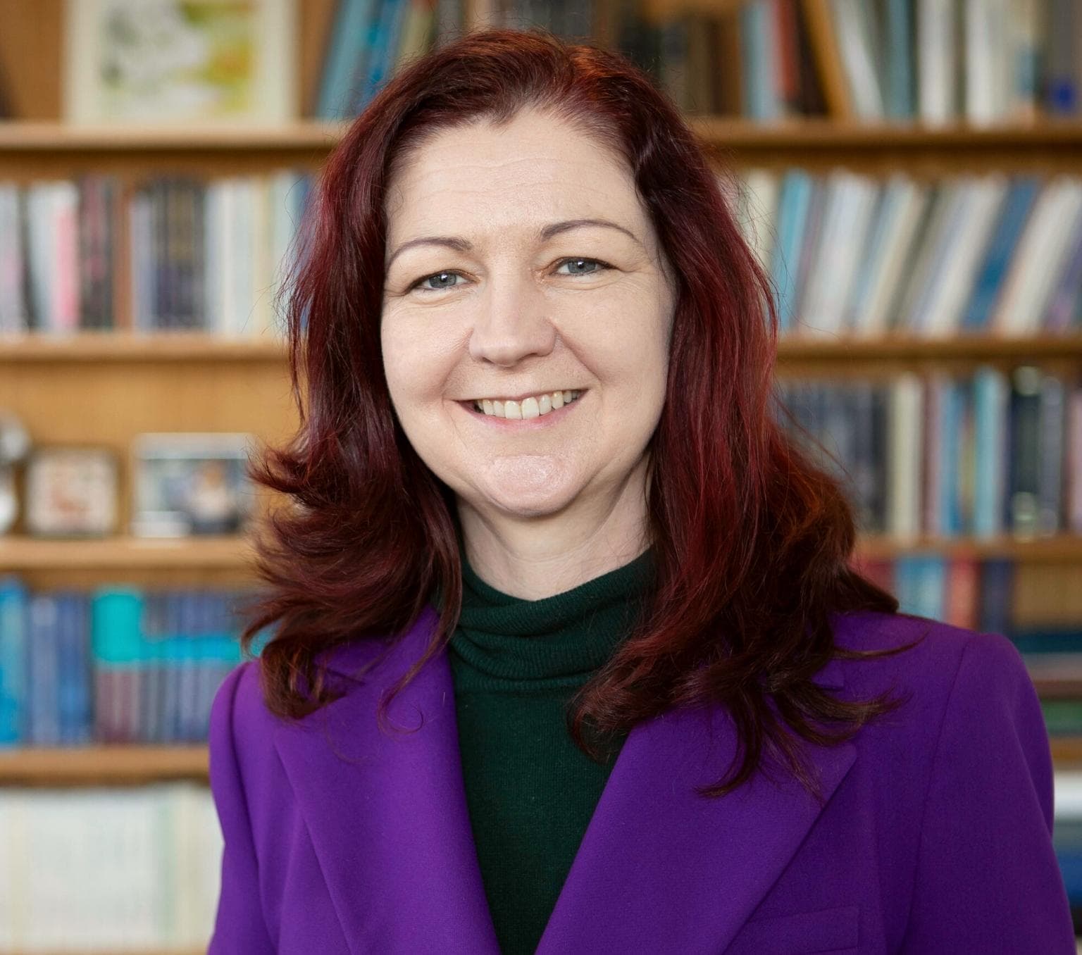 Lisa has dark red hair and is wearing a purple blazer. She is smiling at the camera