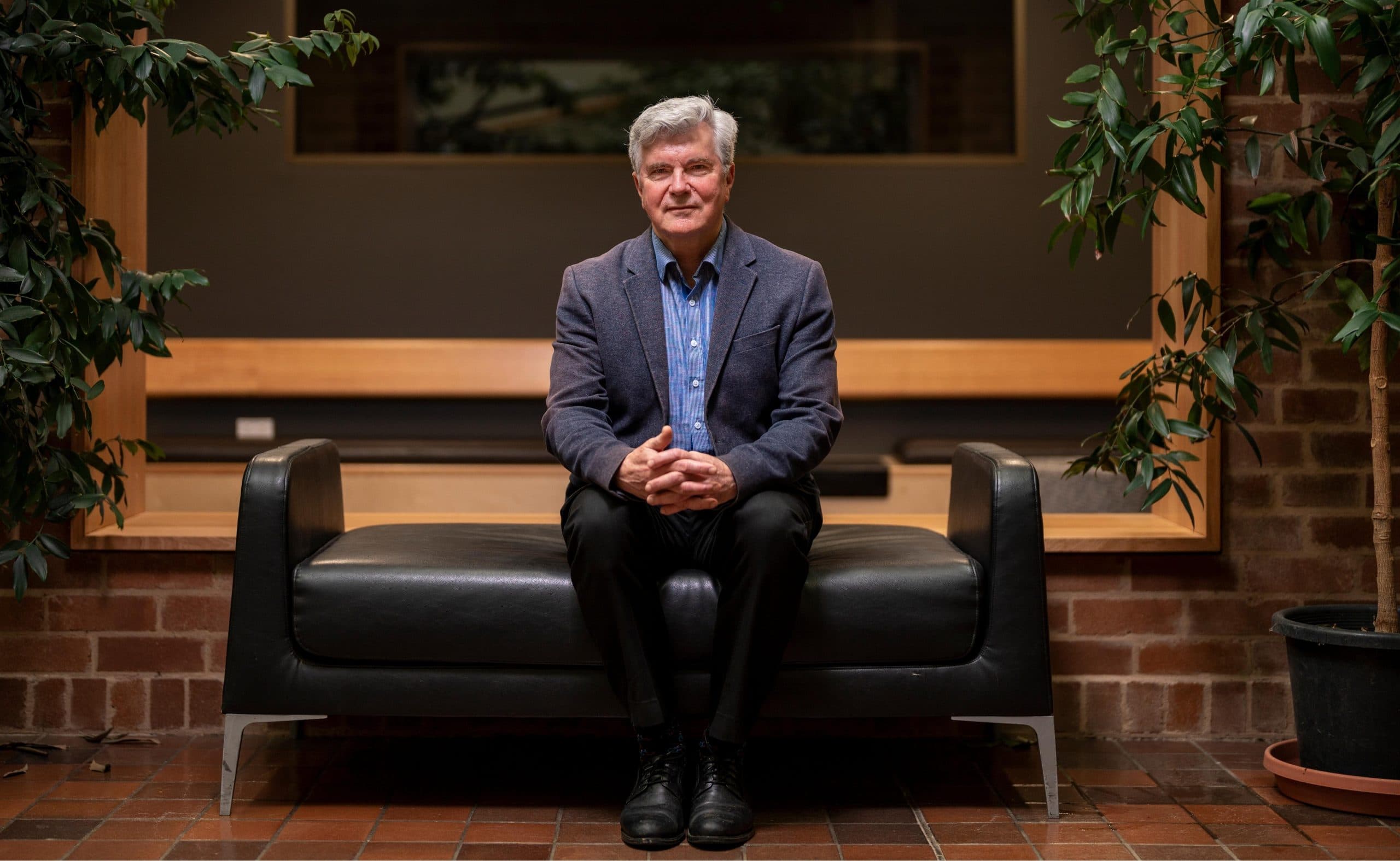 Mark Howden is sitting with his hands clasped on this knees. He is wearing a dark suit and blue collard shirt. He is sitting on a black leather sofa.