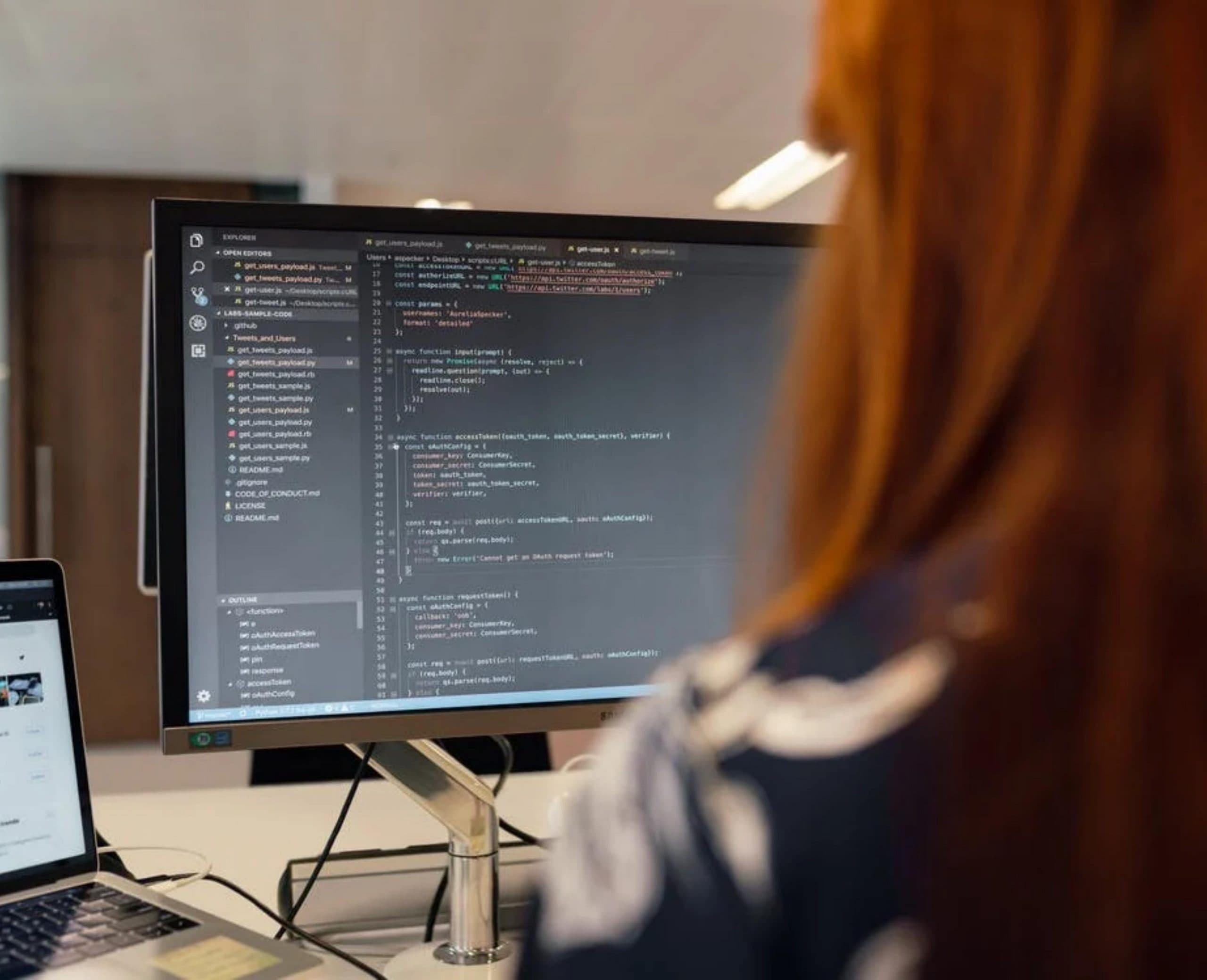 Close up on the back of a woman's head and a computer screen. The woman has red hair and the computer screen has a black screen with colored lines of text.