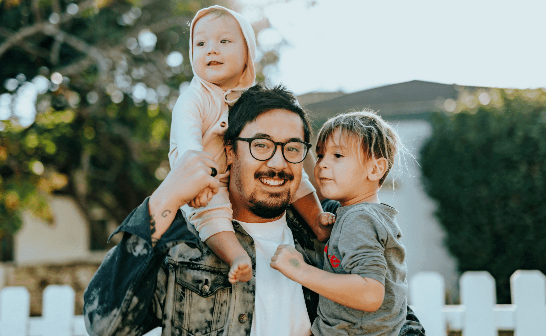 A smiling beared man in glasses has a baby in a onesie on his shoulders and has a little boy wearing a hoodie hoisted on his left side.