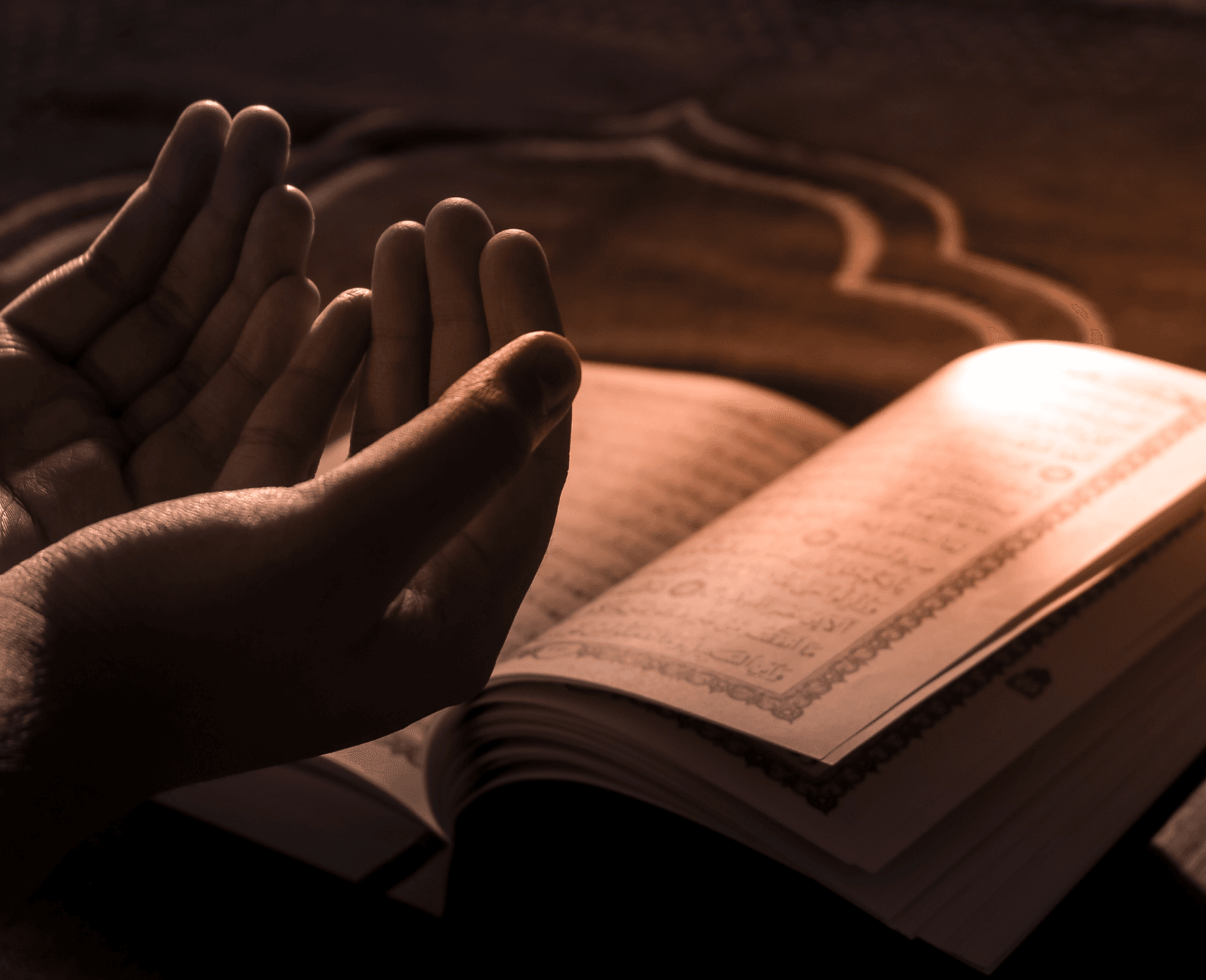 Two hands are cupped facing upwards in front of an open book with Arabic text on the page.