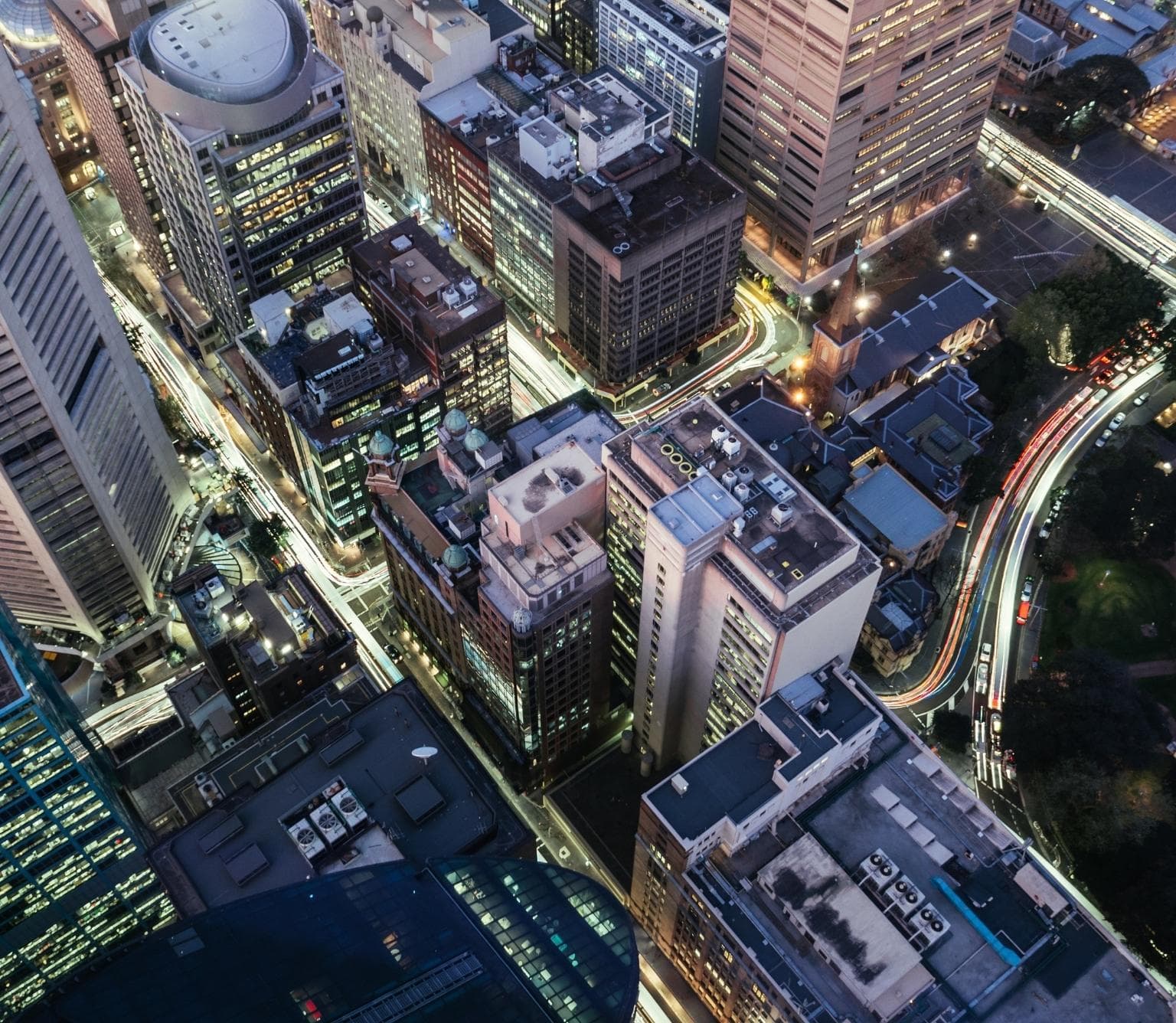 aerial perspective of skyscrapers at dusk in the Sydney CBD. The buildings are mostly shades of grey and brown. flashes of lights are visible on the road from cars driving.