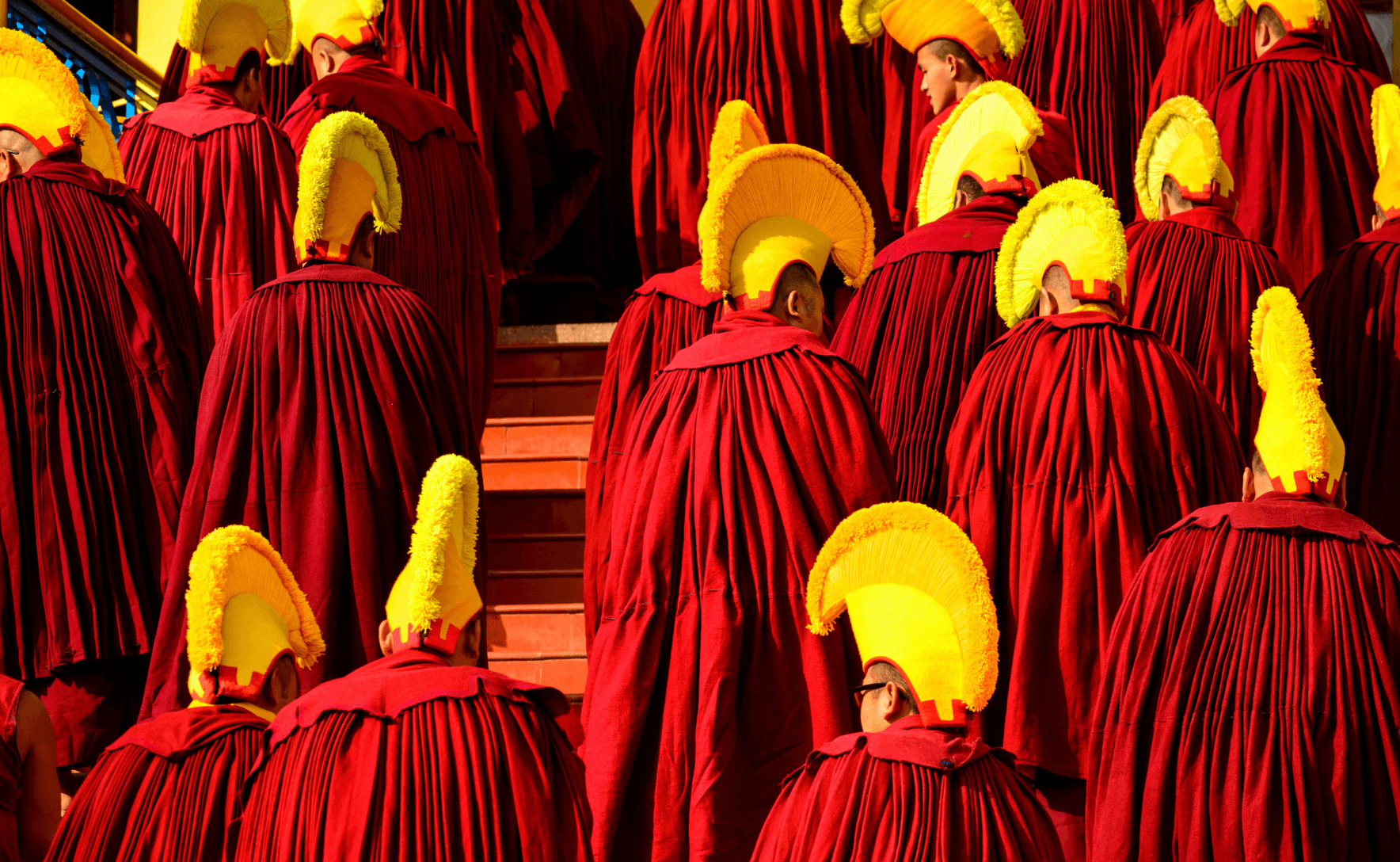 Monks dressed in red robes and yellow head dresses climb stairs outside a monastery.