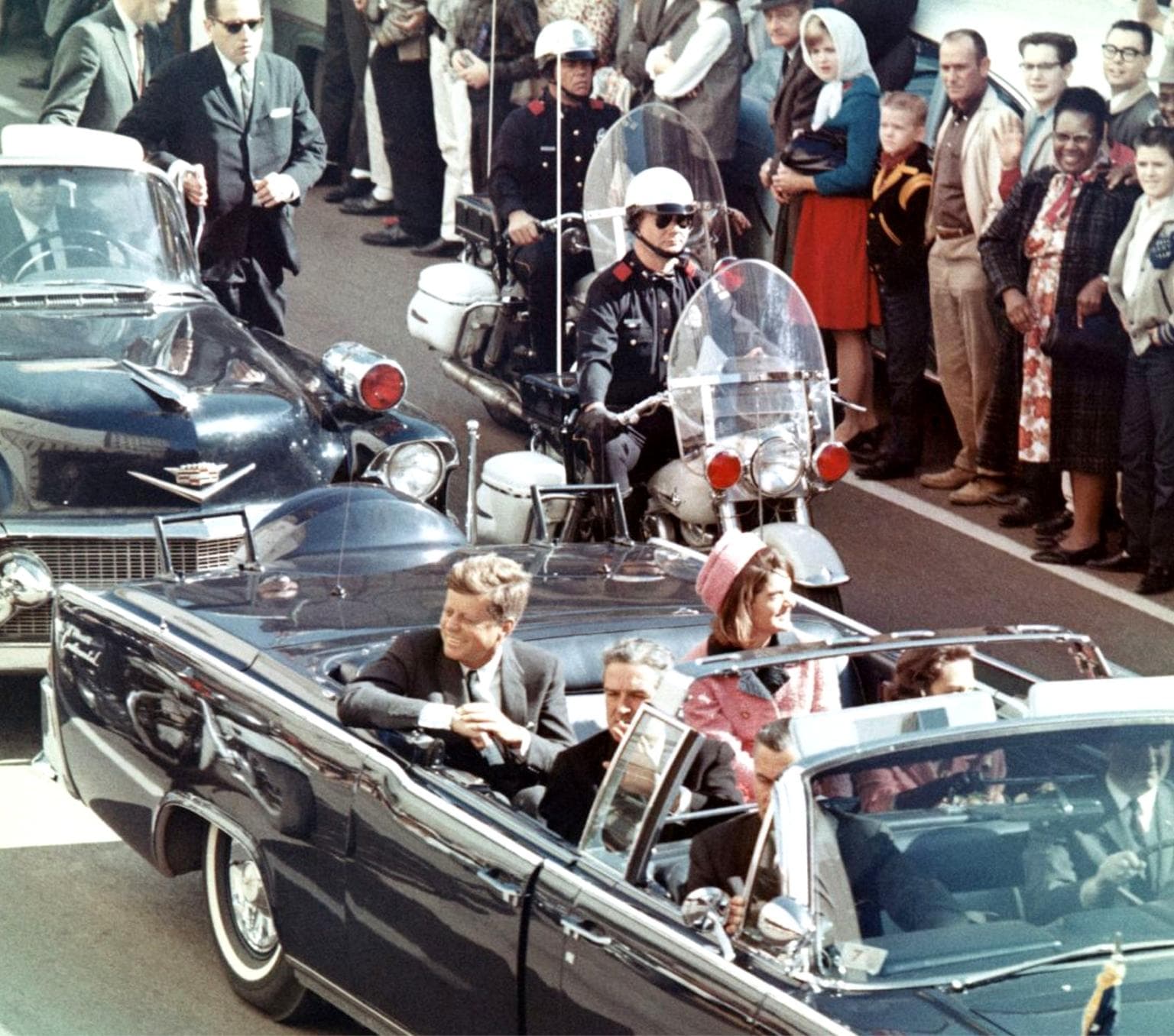 President John F Kennedy sits in the back seat of a convertible limousine. A policeman on a motorbike follows the car. The street is lined with people.