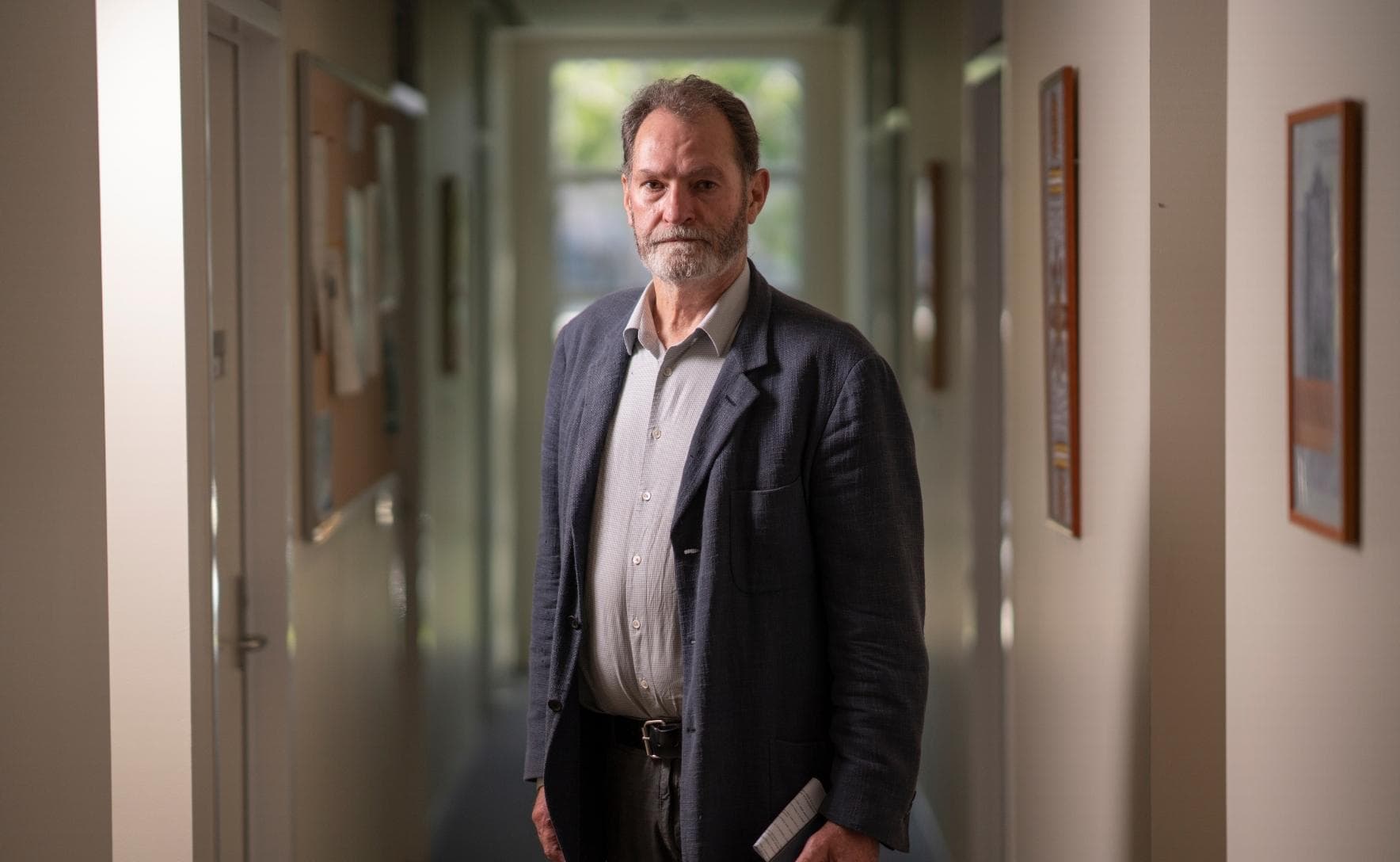 Roderic is wearing a dark blue suit jacket and light grey collared shirt. He is standing in a corridor and holding a piece of paper and a phone.