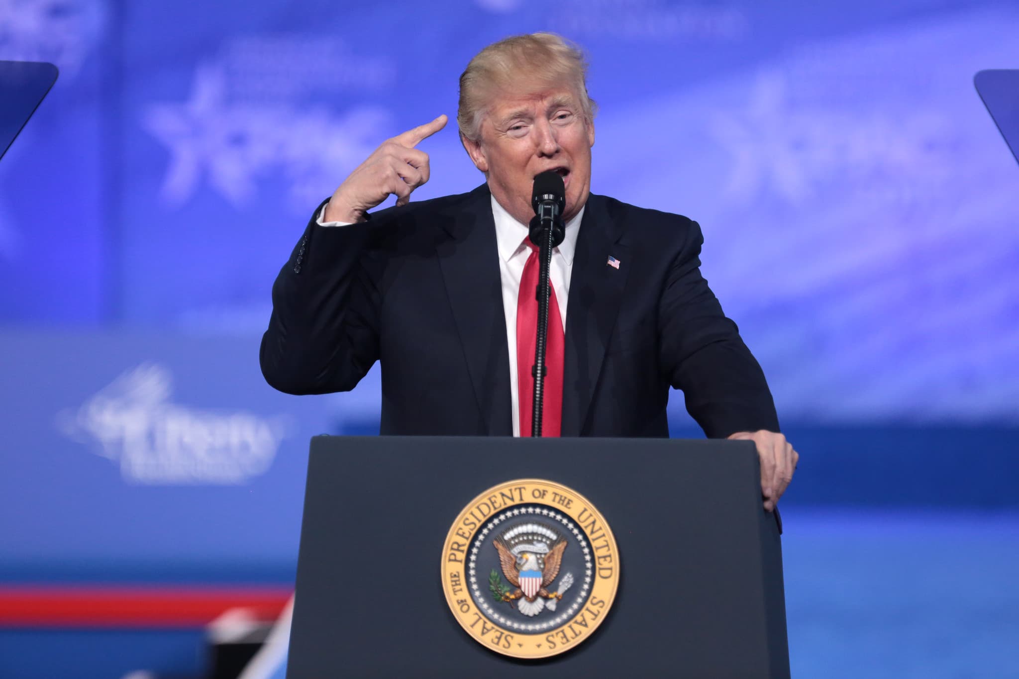 Donald Trump stands at a lecturn with a US president emblem. He is wearing a black suit and read tie. He is mid-speech and is pointing at his head