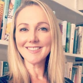 Professor Kate Mitchell smiles in front of a book case. She has blond hair and blue eyes.