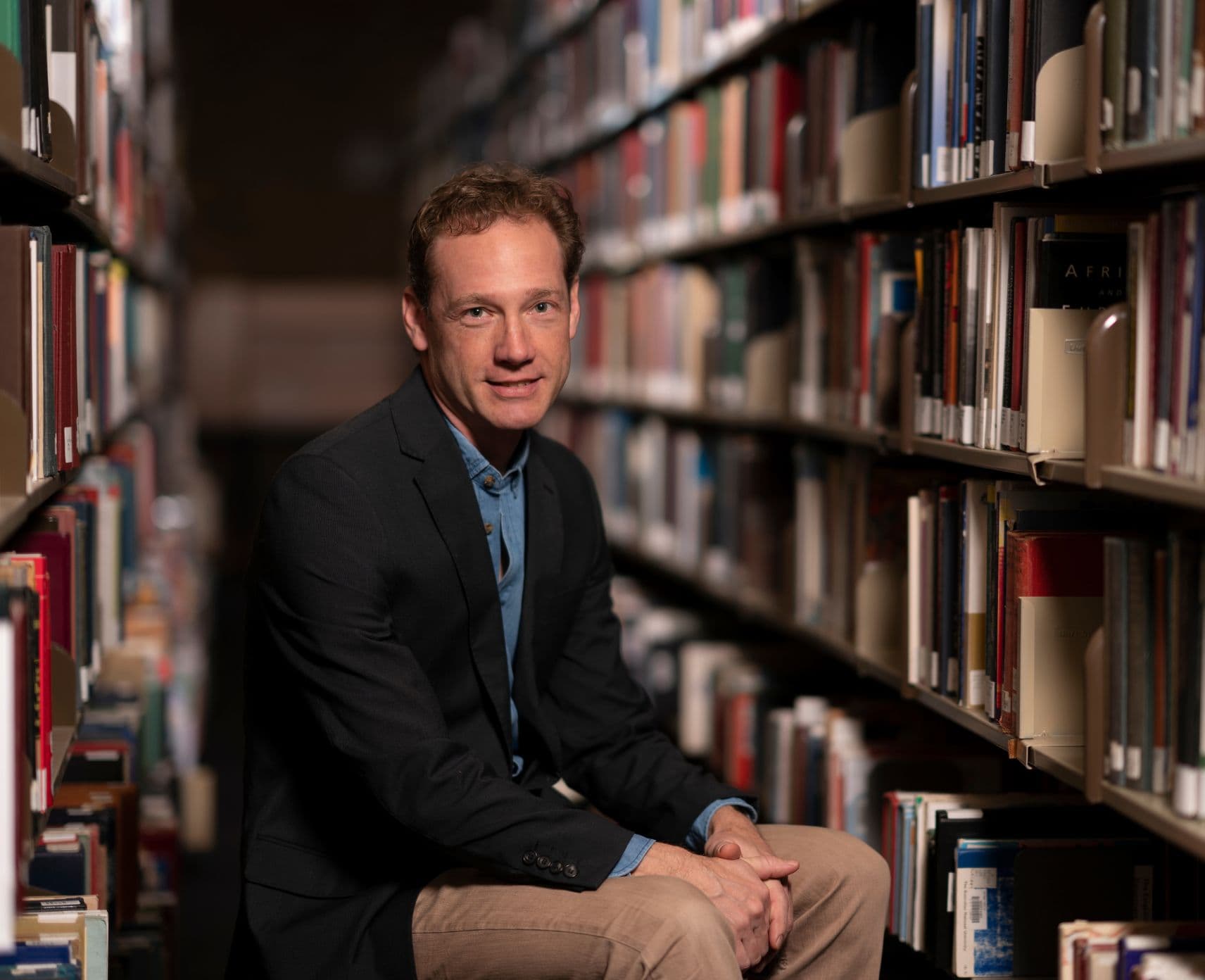 Man in black jacket and tan pants sitting in a row of books