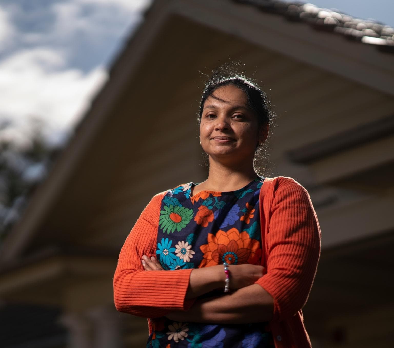 Aparna is wearing a blue dress with orange, blue and green flowers and an orange cardigan. She is looking at the camera with a neutral expression and her arms are folded