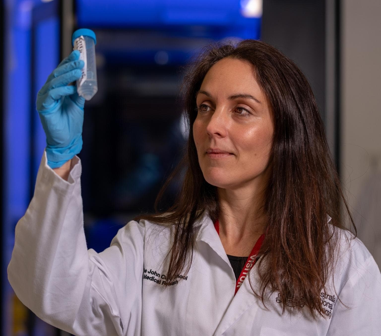 Marian Burr is wearing a white lab coat and has long brown hair. She is tolding up a plastic testube with a blue lid and looking at it