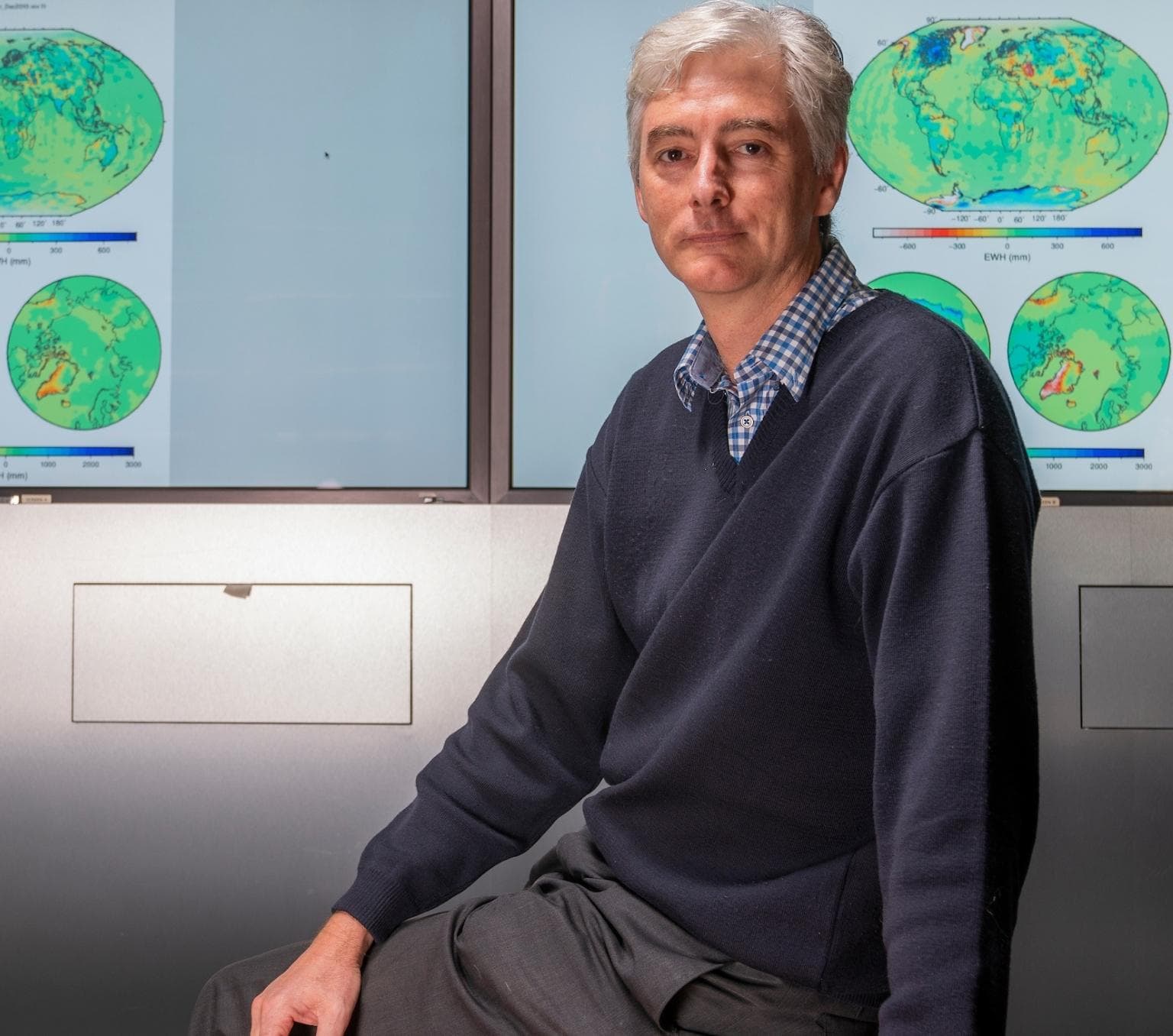 Paul is pictured in a dark blue jumper and grey pants. He is istting in front of a wall with maps of the globe