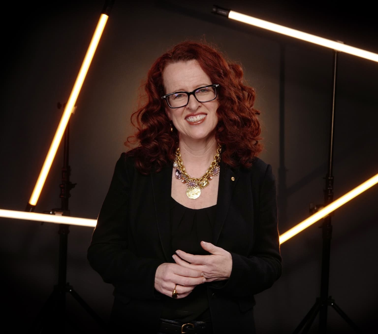 A woman is similing at the camera pictured from the waist up. She is waring a black top and jacket, a bright gold necklace with large jewels and has long curly red hair.