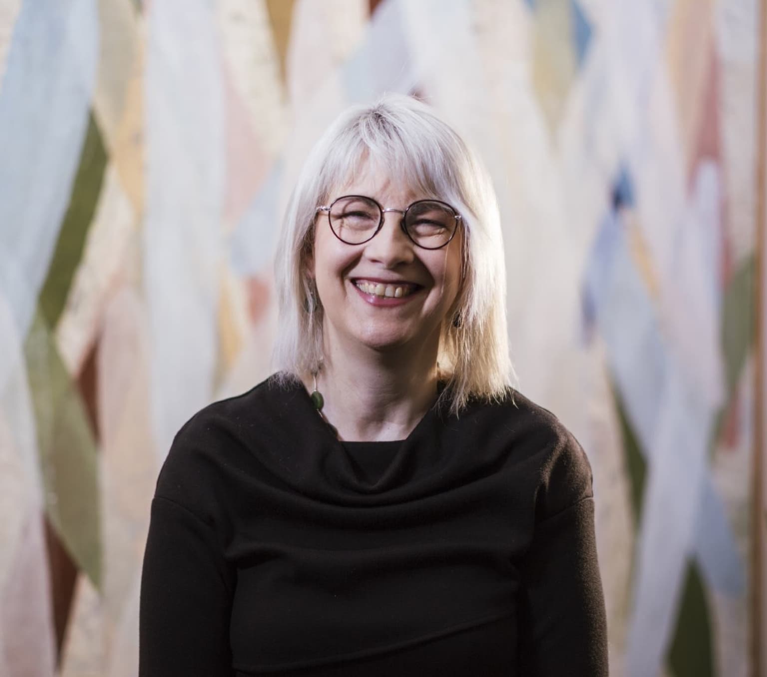 woman with white hair is smiling at the camera. She is wearing glasses and a black top.