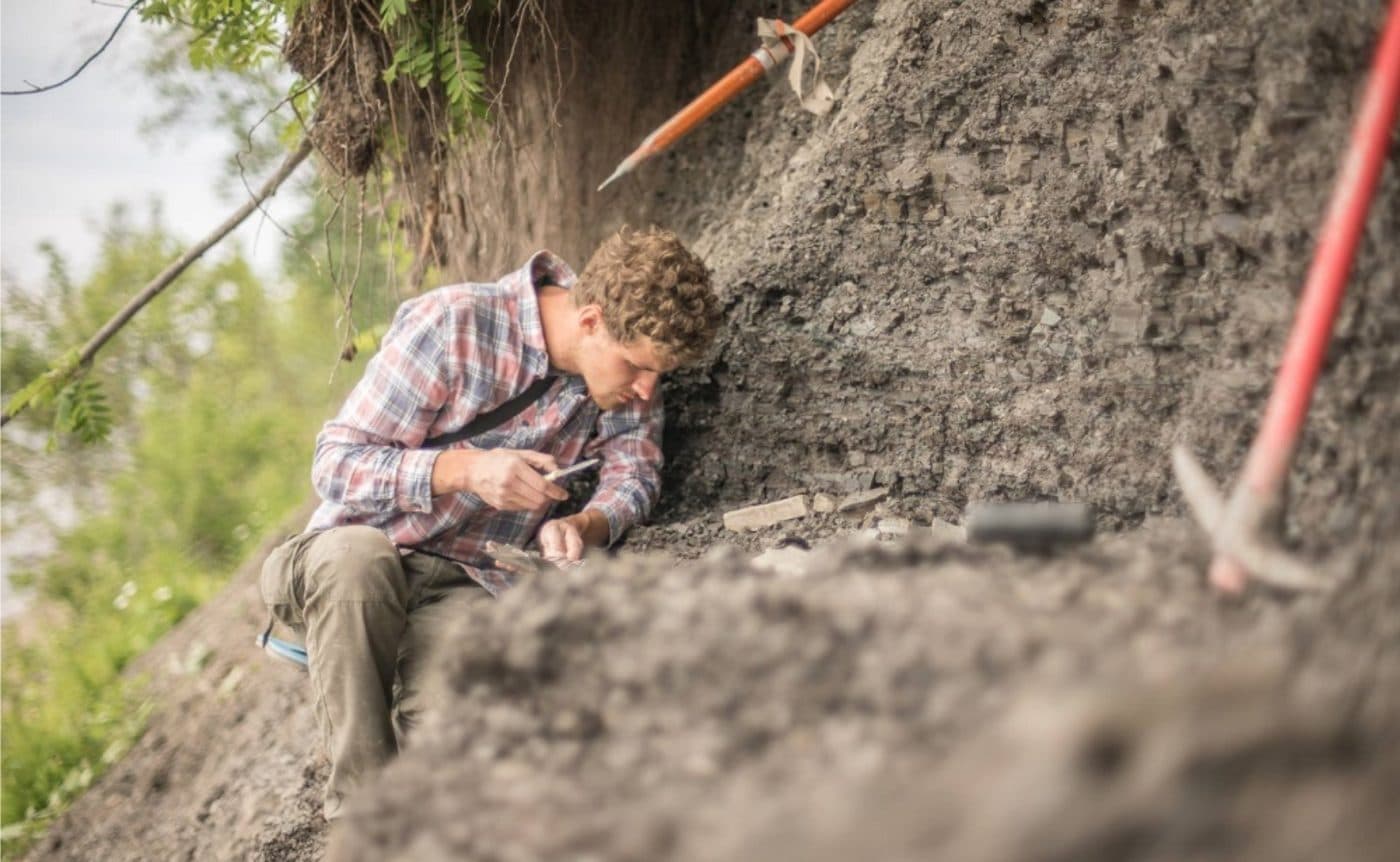 Dr Ilya Bobrovskiy searching for fossils in rock sediment near the White Sea in Russia. He is wearing a pink and blue checkered shirt and has excavation equipment next to him. 