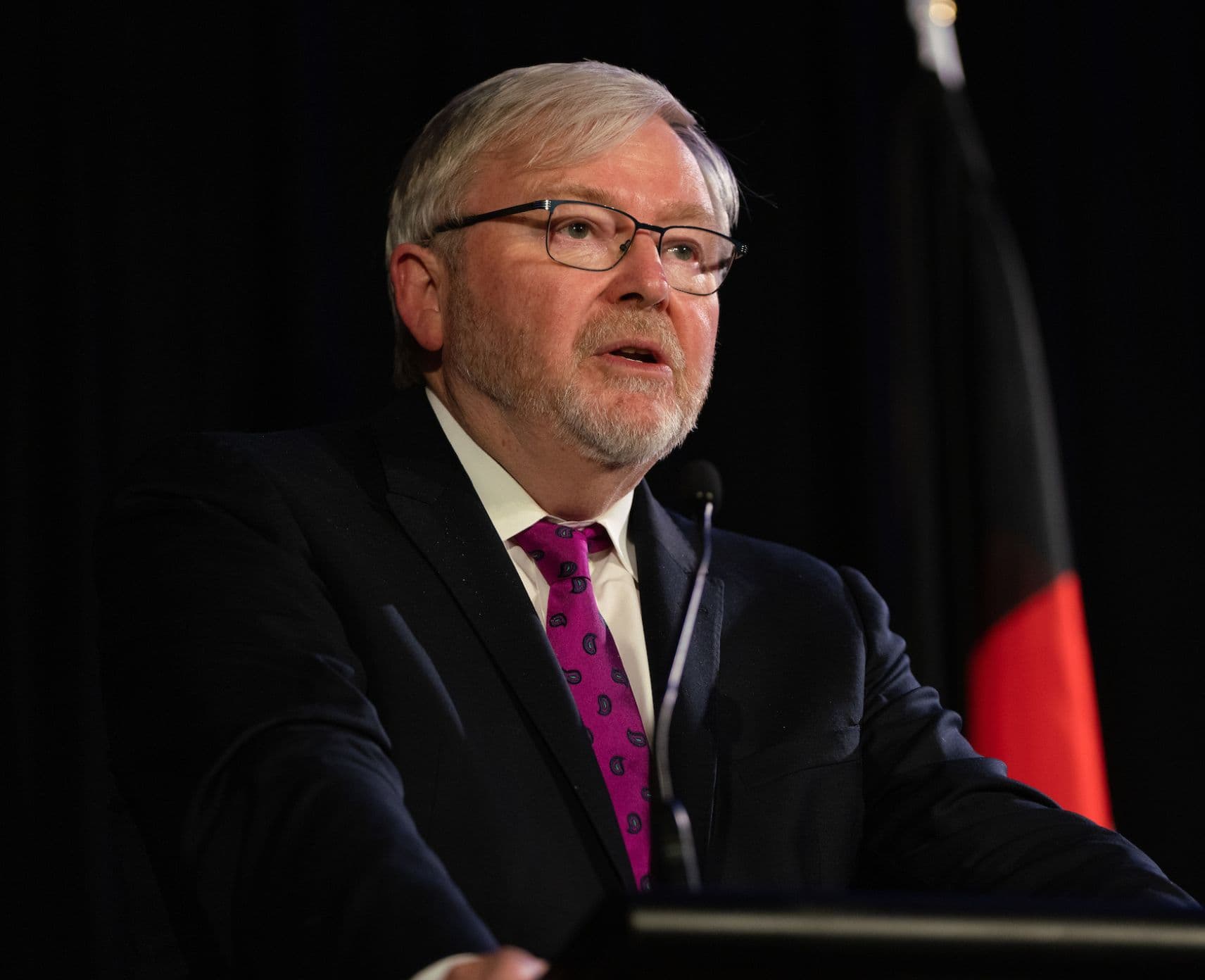 A man with white hair and a white beard wearing glasses and a black suit speaking at a lectern.