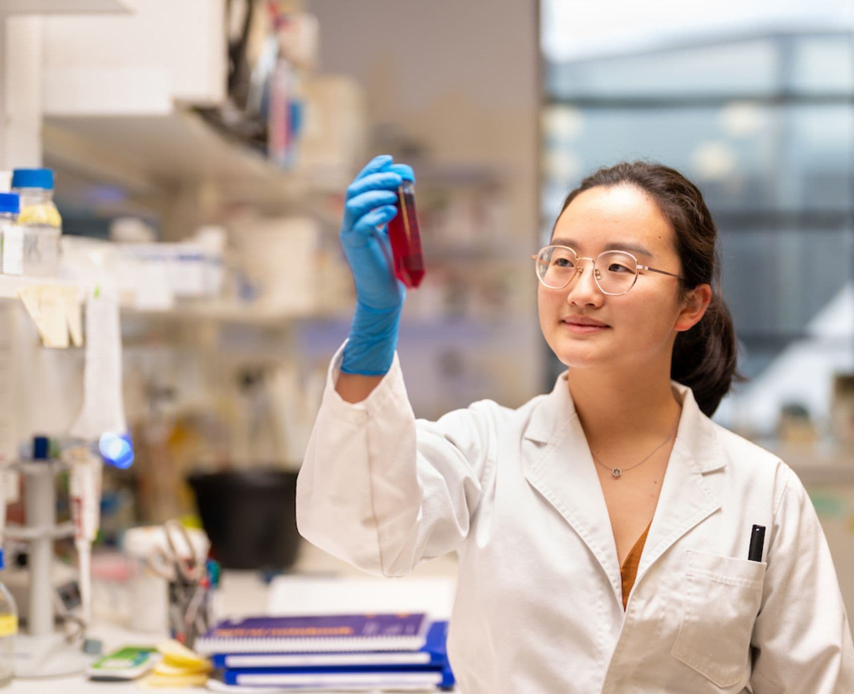 Jennifer Hung is in a lab holding a vial of chemicals. She wears a white lab coat, blue gloves and a pair of spectacles. Her dark hair is in a ponytail.