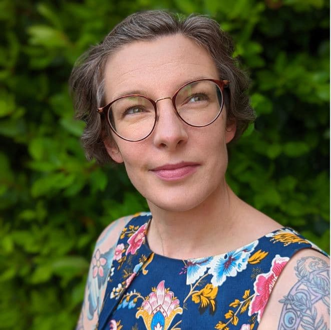 Mary Dahm looks to the side of the camera. She has short brown hair, round glasses and a floral sleeveless dress. She has tattoos on her upper arms.