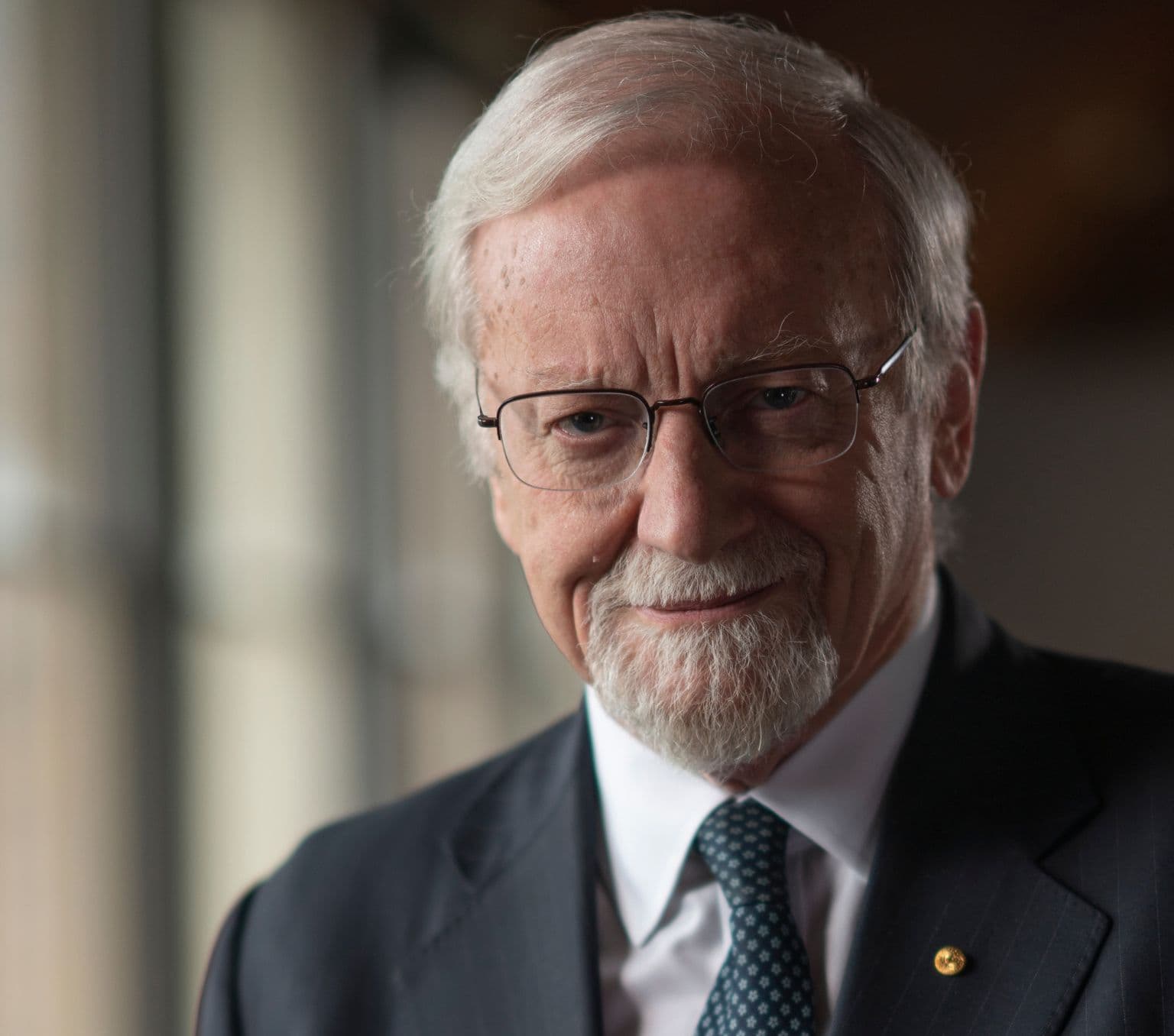 A man with white hair wearing glasses a blue suit jacket looks into the camera.