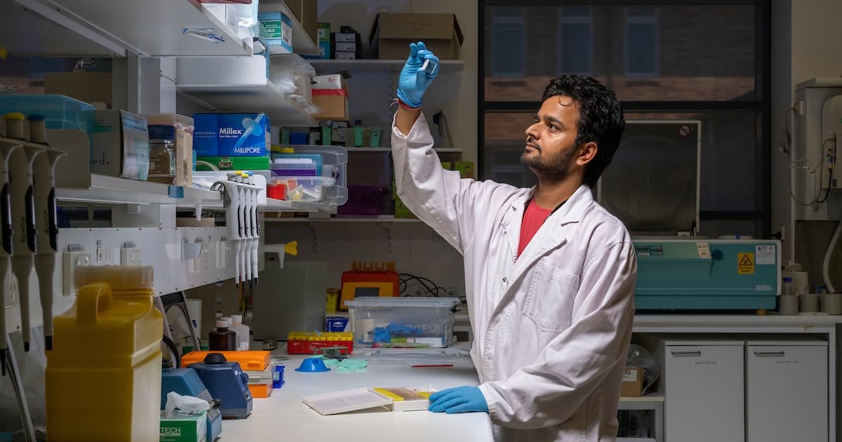 Dr Abhimanu Pandey inspects a test tube containing an unknown substance. He is wearing a white lab coat with a red t-shirt underneath.