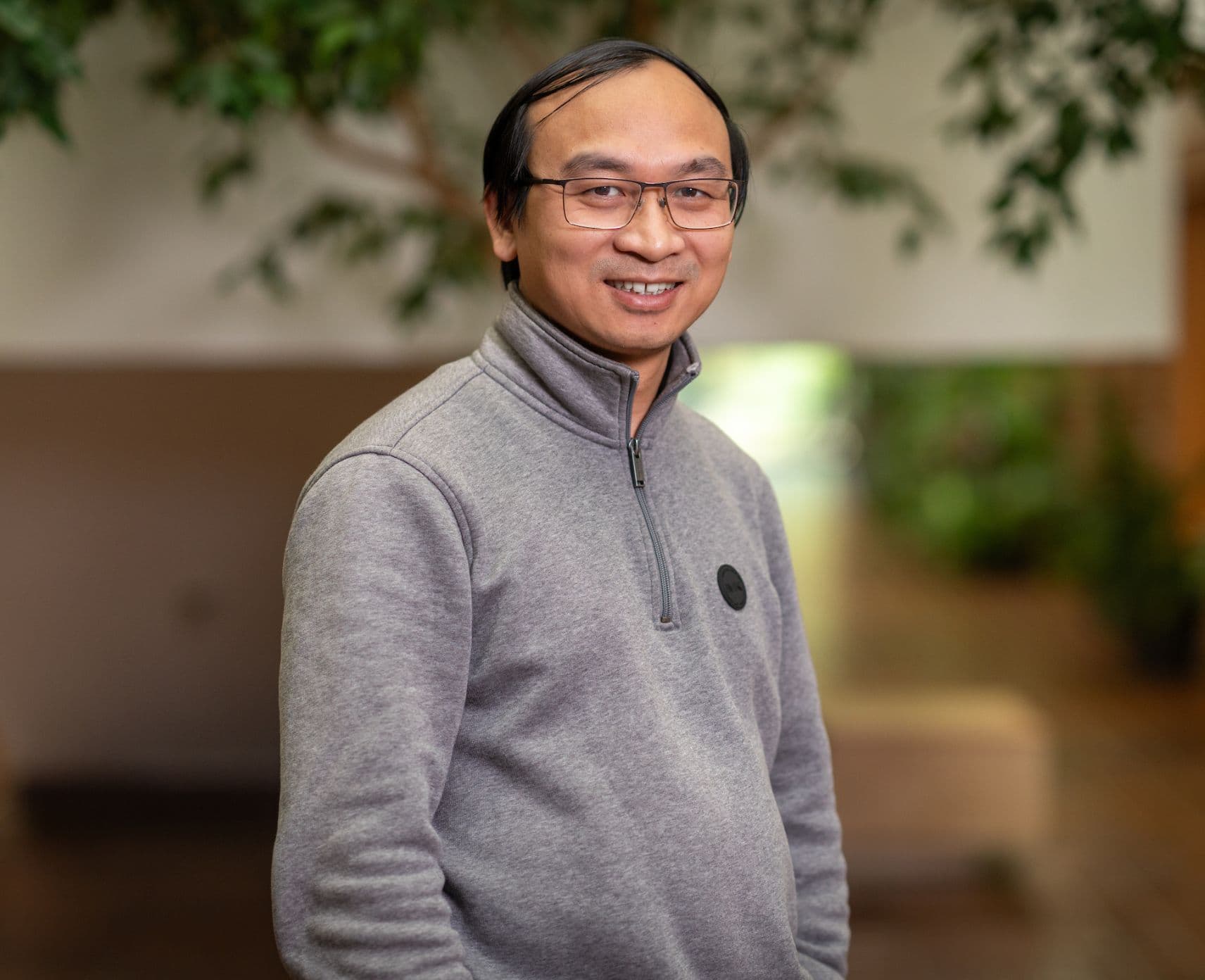 Close-up of Tai. He is smiling and facing the camera. He is wearing a grey jumper and glasses.