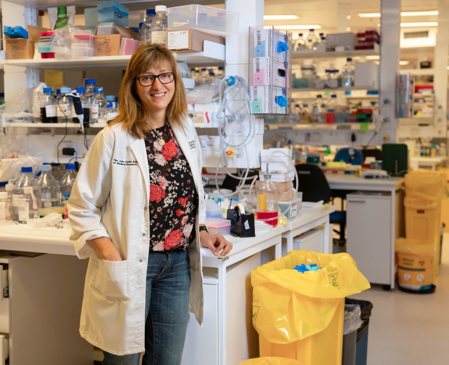 Dr Vicki Athanasopoulos is in a science lab wearing a white lab coat. She is wearing a floral pattern shirt under her lab coat