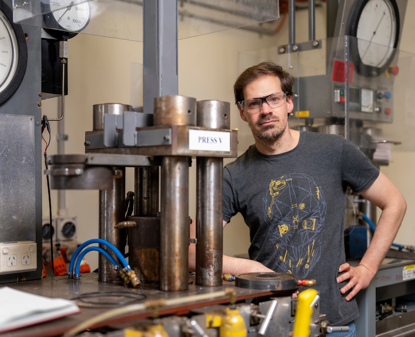 Dr Michael Anenburg is wearing safety goggles and is leaning against a desk next to lab equipment in the ANU Research School of Earth Sciences