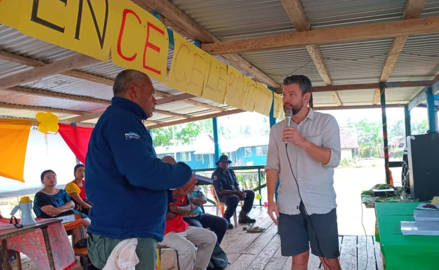 Ben Shaw holds a microphone and looks towards the president of a local level government. They are in an open structure outdoors with a large homemade banner in the background