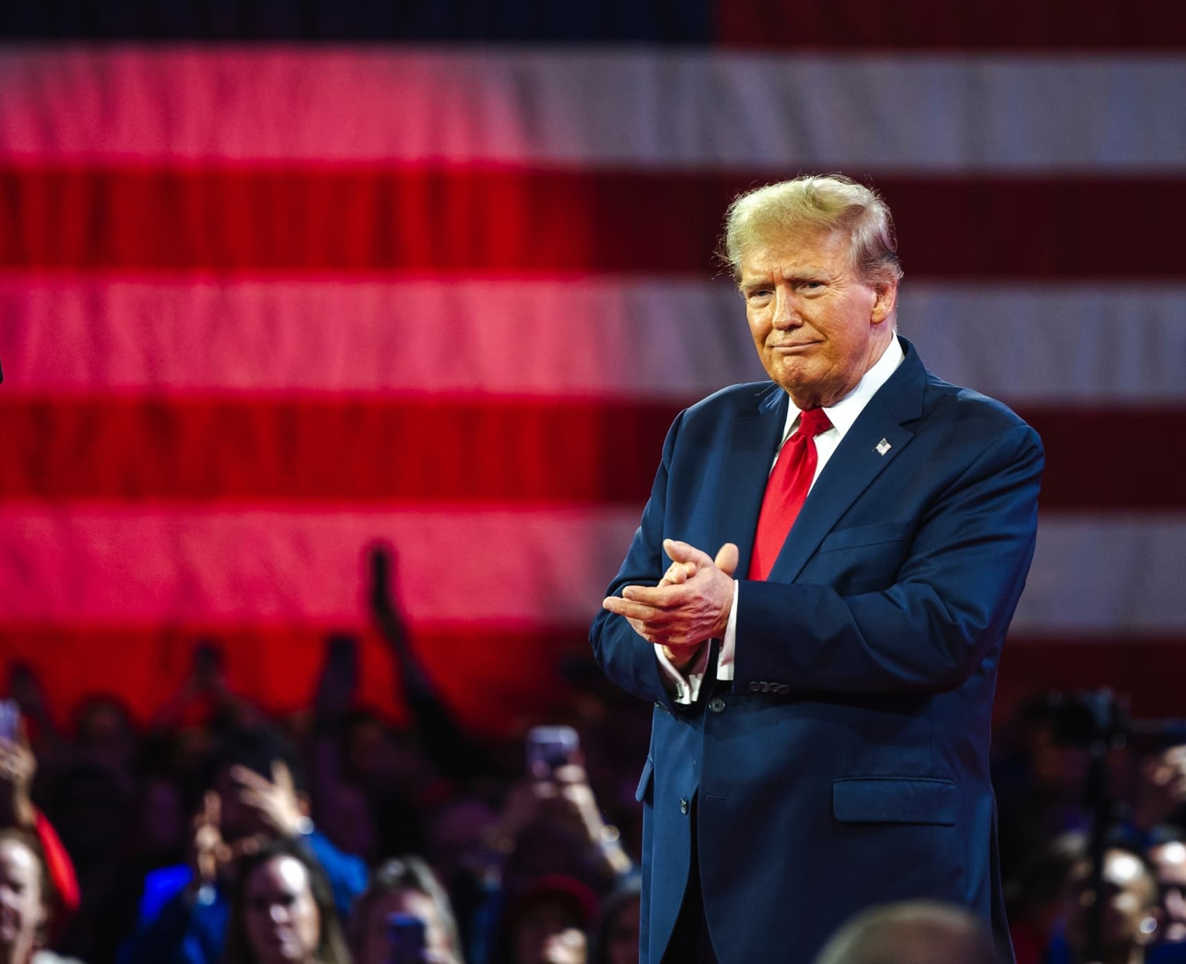 Donald Trump with hands clasped in front of rally crowd. A large American flag with a red spotlight in background.