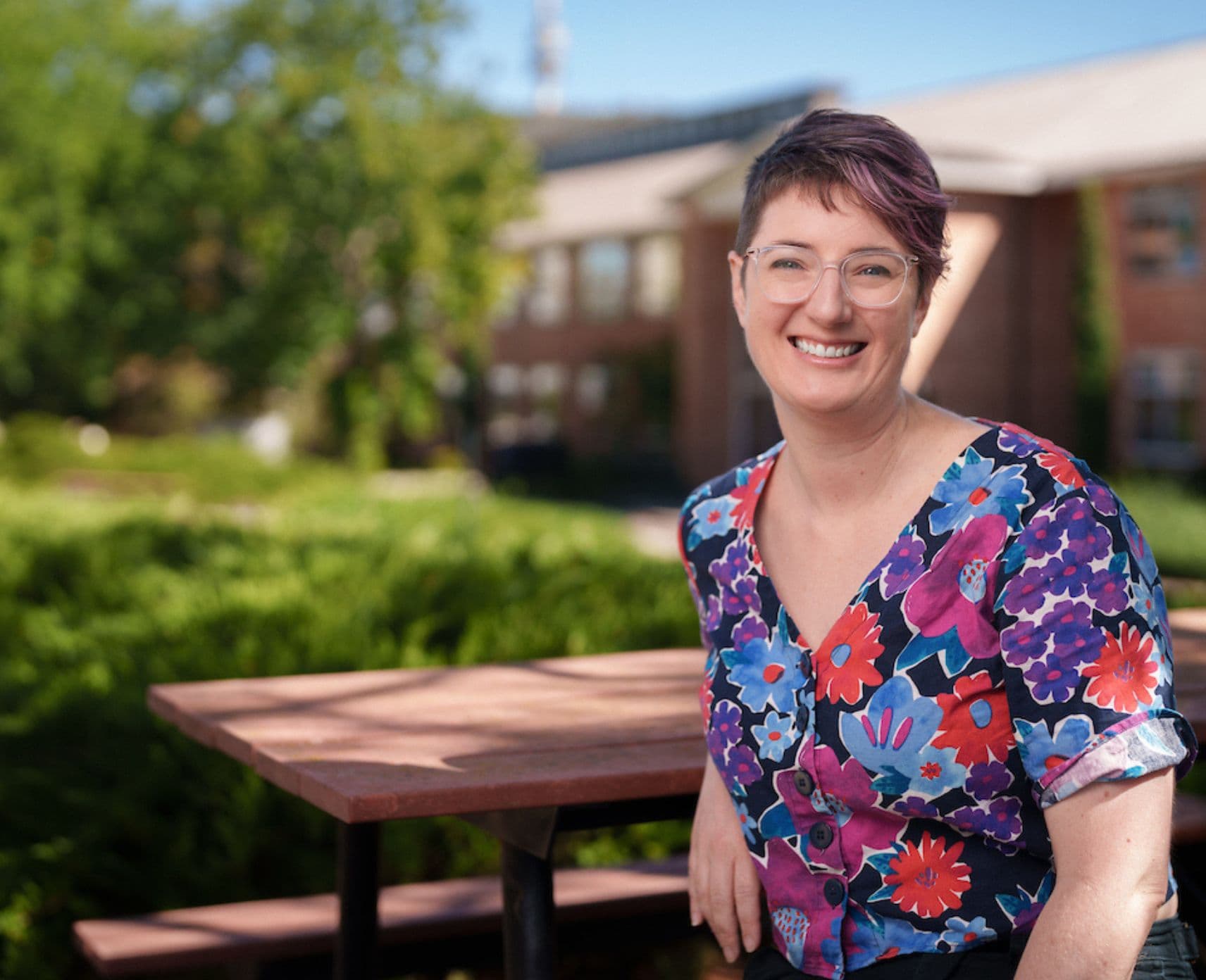 PhD scholar Kai Hodgkin smiles at the camera. She has short pink hair, glasses and wears a colourful floral top.