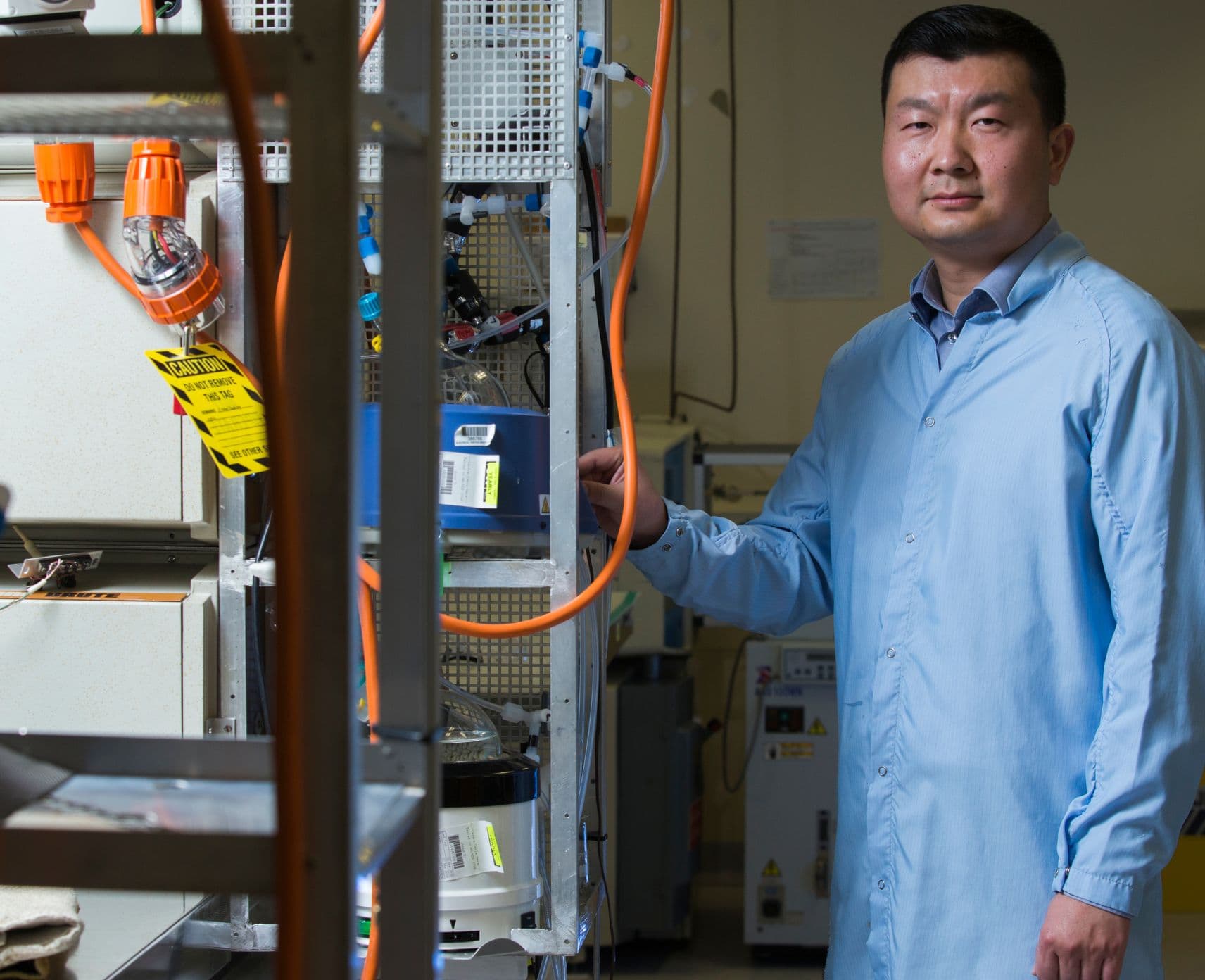Bin Lu standing next to equipment in his lab, wearing a blue shirt