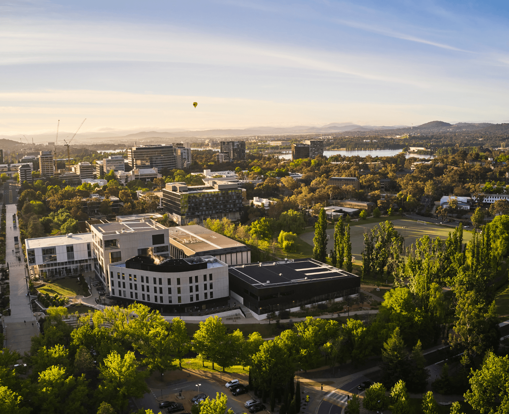 An aerial photograph of the ANU campus, Canberra central business district and Lake Burley Griffin, with a hot air baloon in the distance.