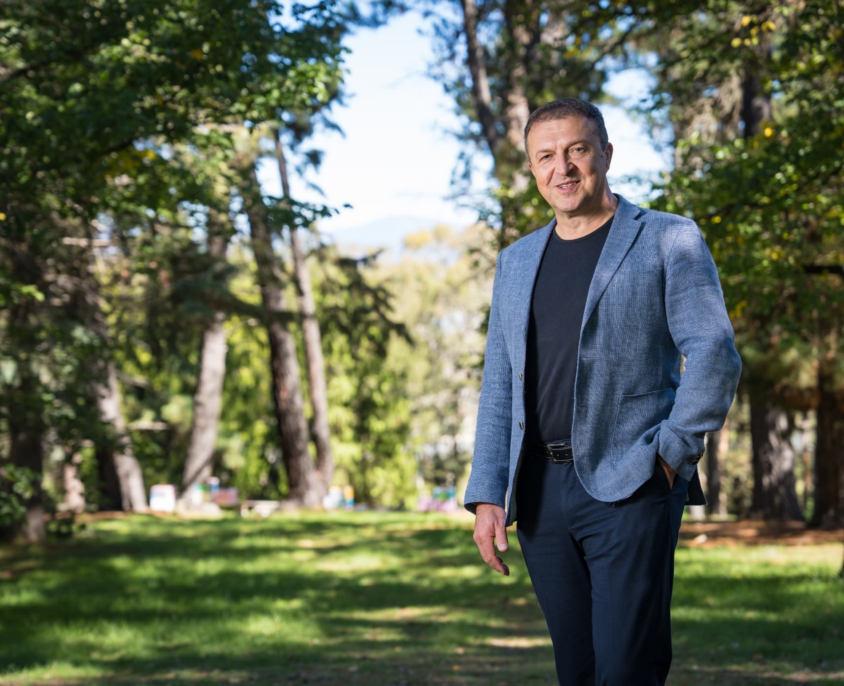Professor Hrvoje Tkalčić is wearing a blue blazer with a black shirt underneath. He is standing outside in front of trees and has one hand in his pocket