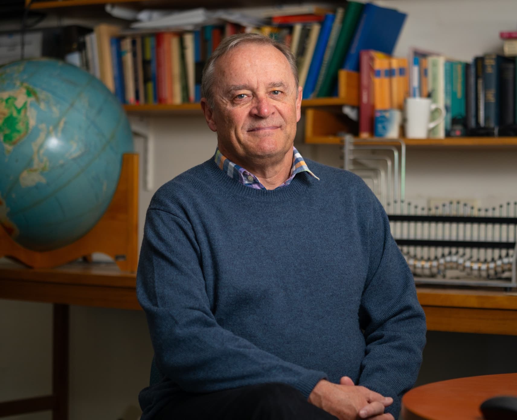 Emeritus Professor Malcolm Sambridge sits on a chair with a globe of the earth behind him. There is also a bookshelf behind him.