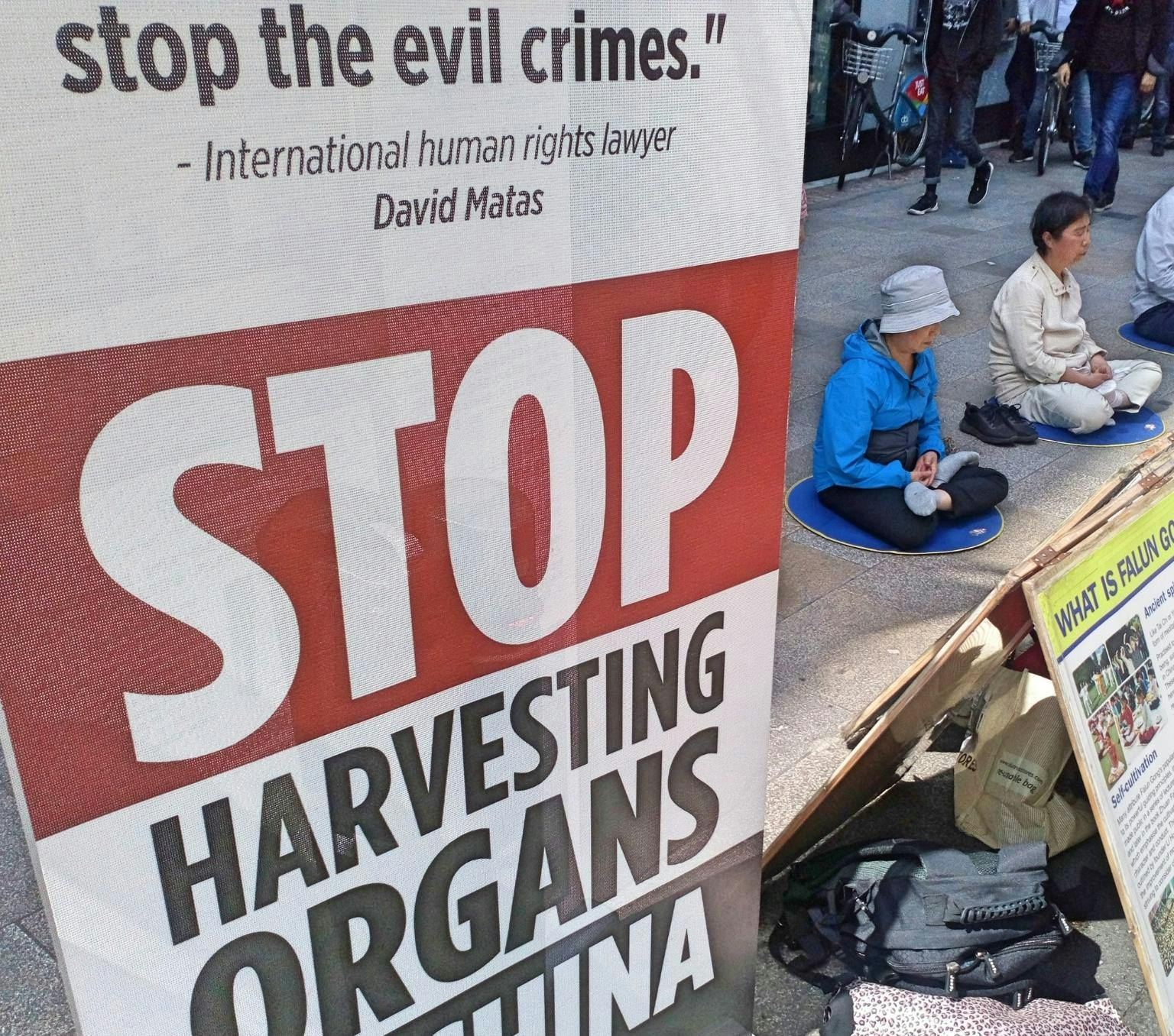 An information stall on organ harvesting in China on display in Dublin, Ireland. There is a sign that reads: 'STOP organ harvesting in China'. There is another sign that reads: what is Falun Gong?' There are people sitting on the floor with their legs crossed in the background.