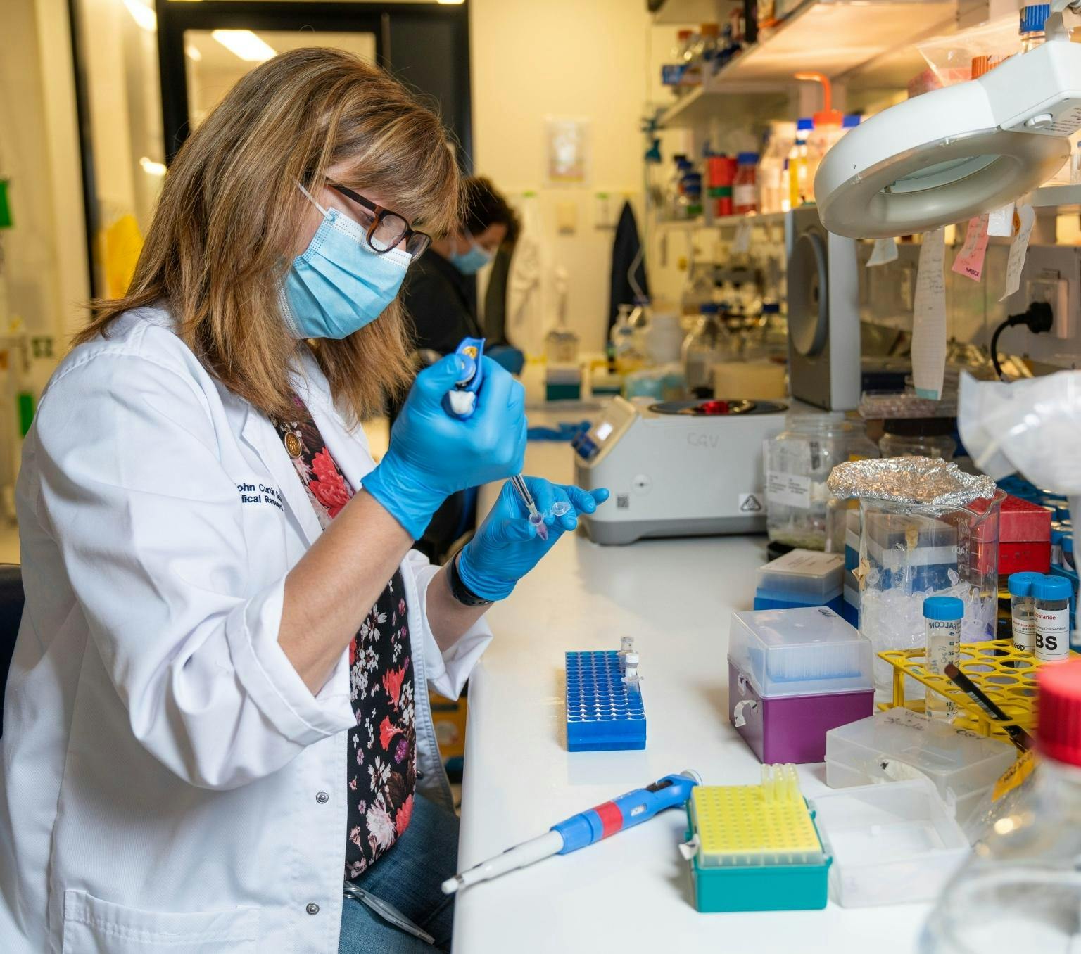 Dr Vicki Athanasopoulos works on lupus research in the lab. She is wearing a white lab coat and blue medical gloves. She is using a measurement device to carefully inject substances into test tubes.
