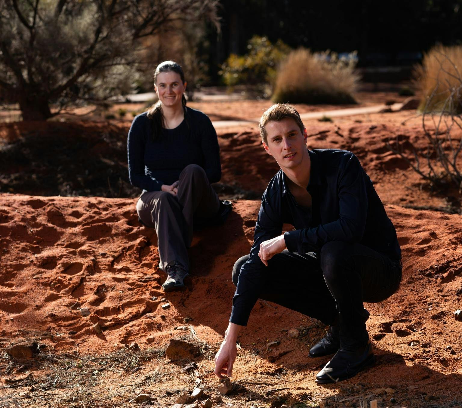 Dr Emma Tucker, wearing dark clothes, sits on a mound of red dirt. Dr Lex van Loon, also wearing dark clothing, squats in front of her, touching a rock on the ground. Behind them is red dirt and shrubs.