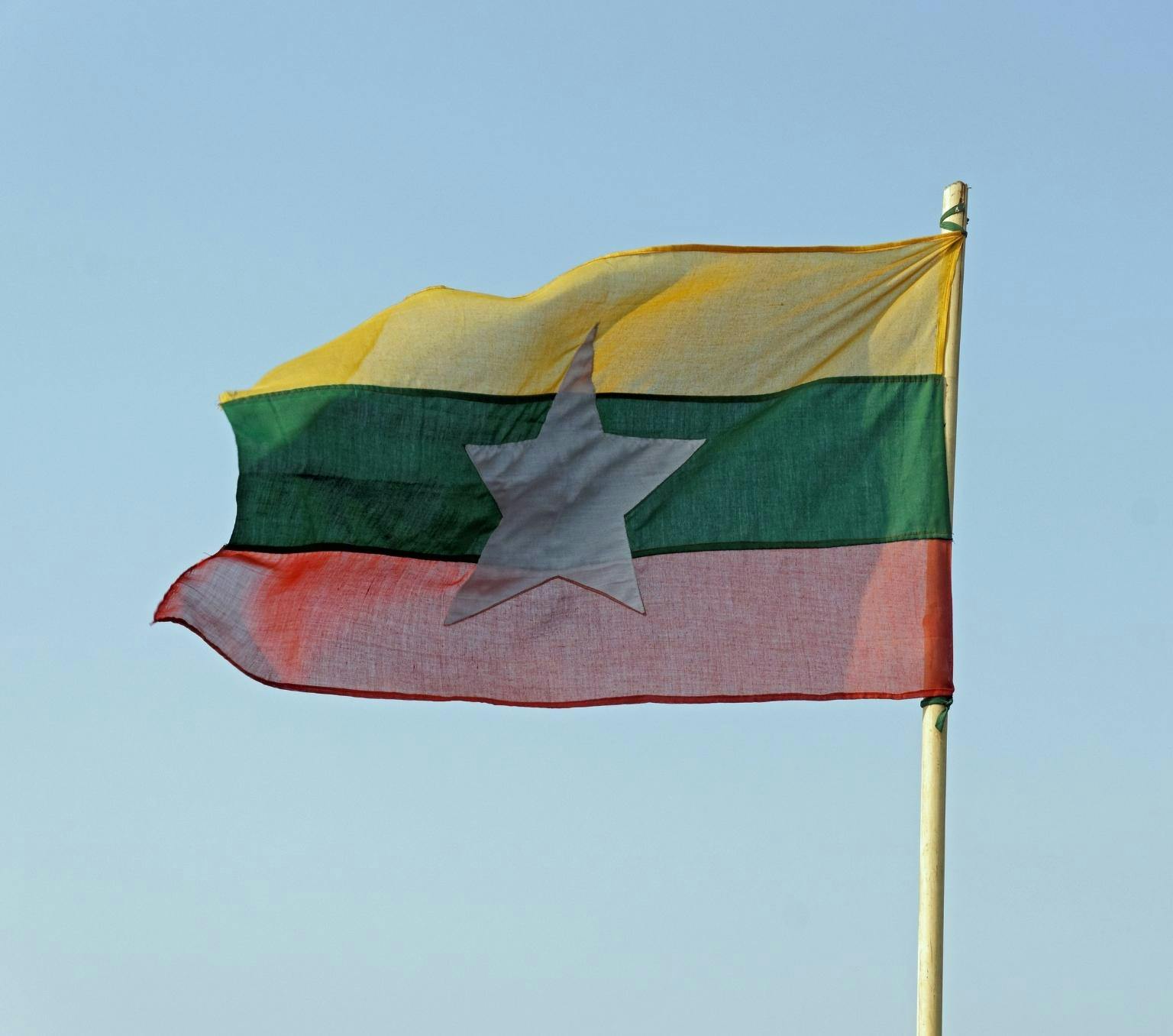 Myanmar flag which has a yellow, green and red horizontal stripe with a five pointed white star in the centre is pictured flying on a flag pole in front of a blue sky