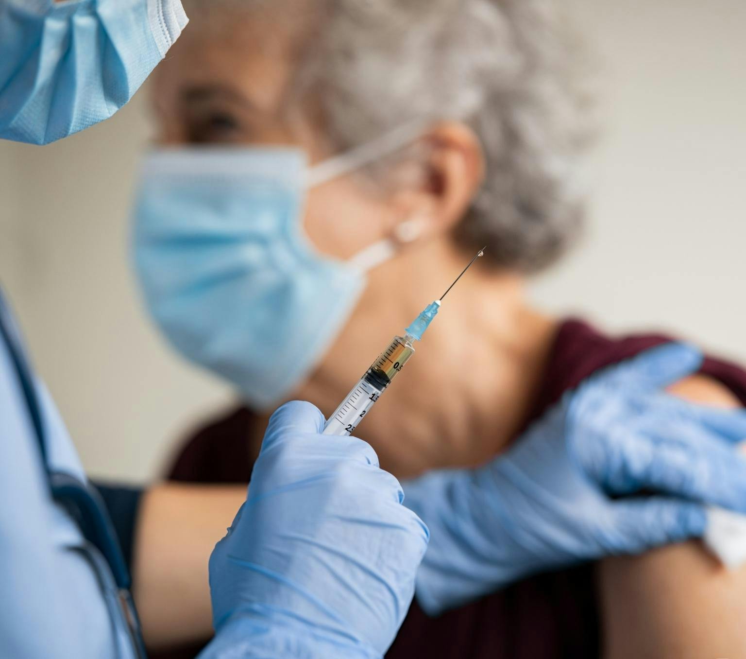 A person wearing blue gloves holds a needle in their right hand. Their left hand touches the bare arm of a woman, who is blurred in the background.