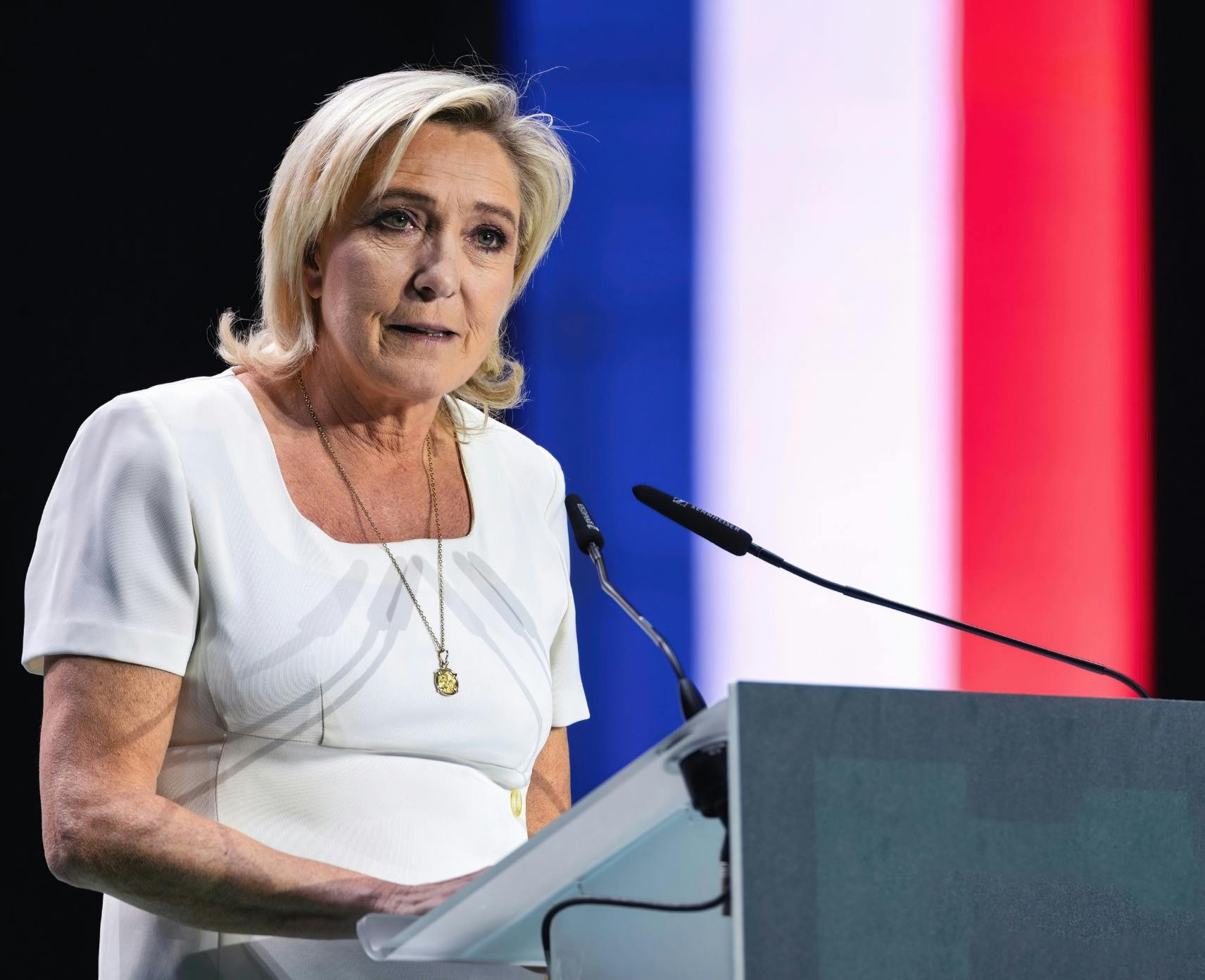 Marine Le Pen, leader of the far-right National Rally party in France. She is standing behind a podium and there is a French flag in the background.
