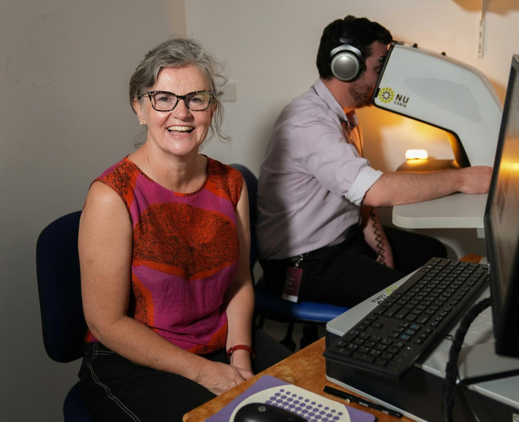 Sarah Mann smiles as man behind is concealed by her machine to measure the emotional power of singing