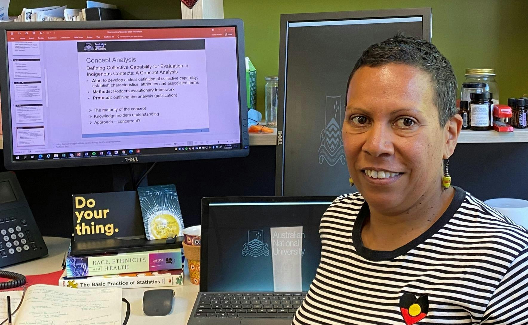 Bobby is wearing a black and white striped top with an aboriginal flag in a love heart shape on her left chest. She is sitting at a desk and smiling
