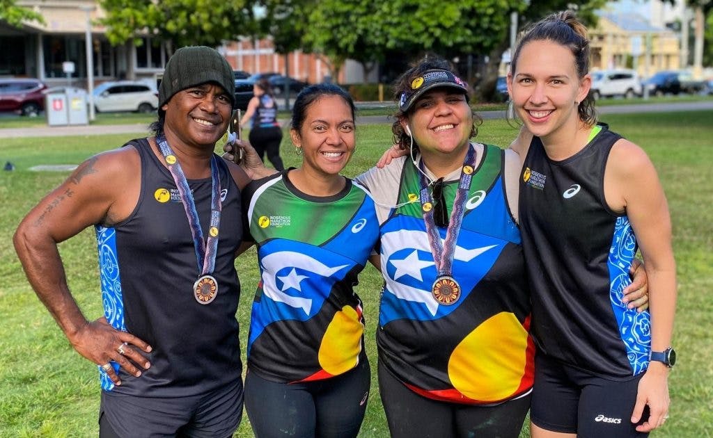 a man and three women are standing in a row and smiling. They are wearing athletic clothing and jerseys. The two women in the centre are wearing jerseys with the Aboriginal flag and Torres Strait Island flag on them