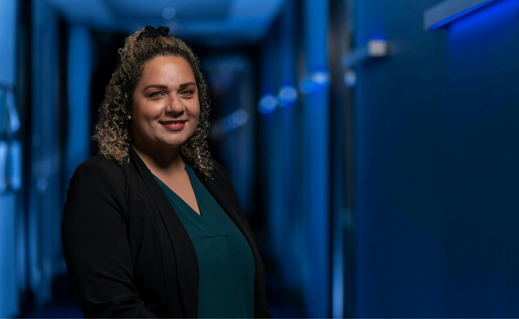 Lisa is wearing a black jacket and dark green blouse. She has dark curly hair and is smiling at the camera. A dark corridor lit with blue lights is in the background