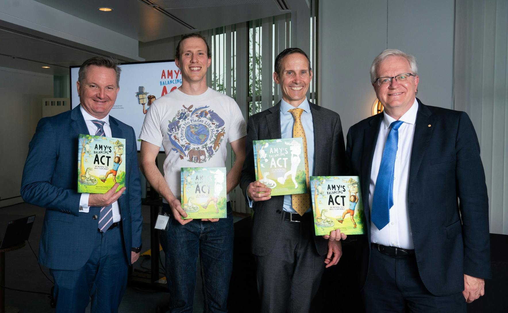 Four men standing in a row and smiling while holding the book Amy's balancing act