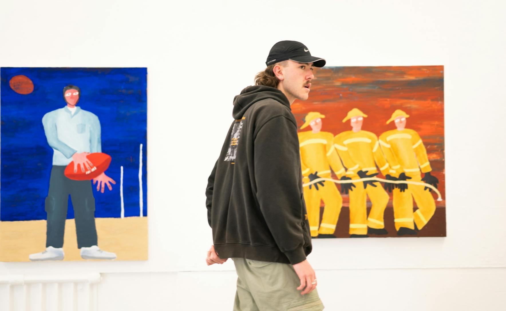 man wearing a capt and black hoodie standing in front of two paintings