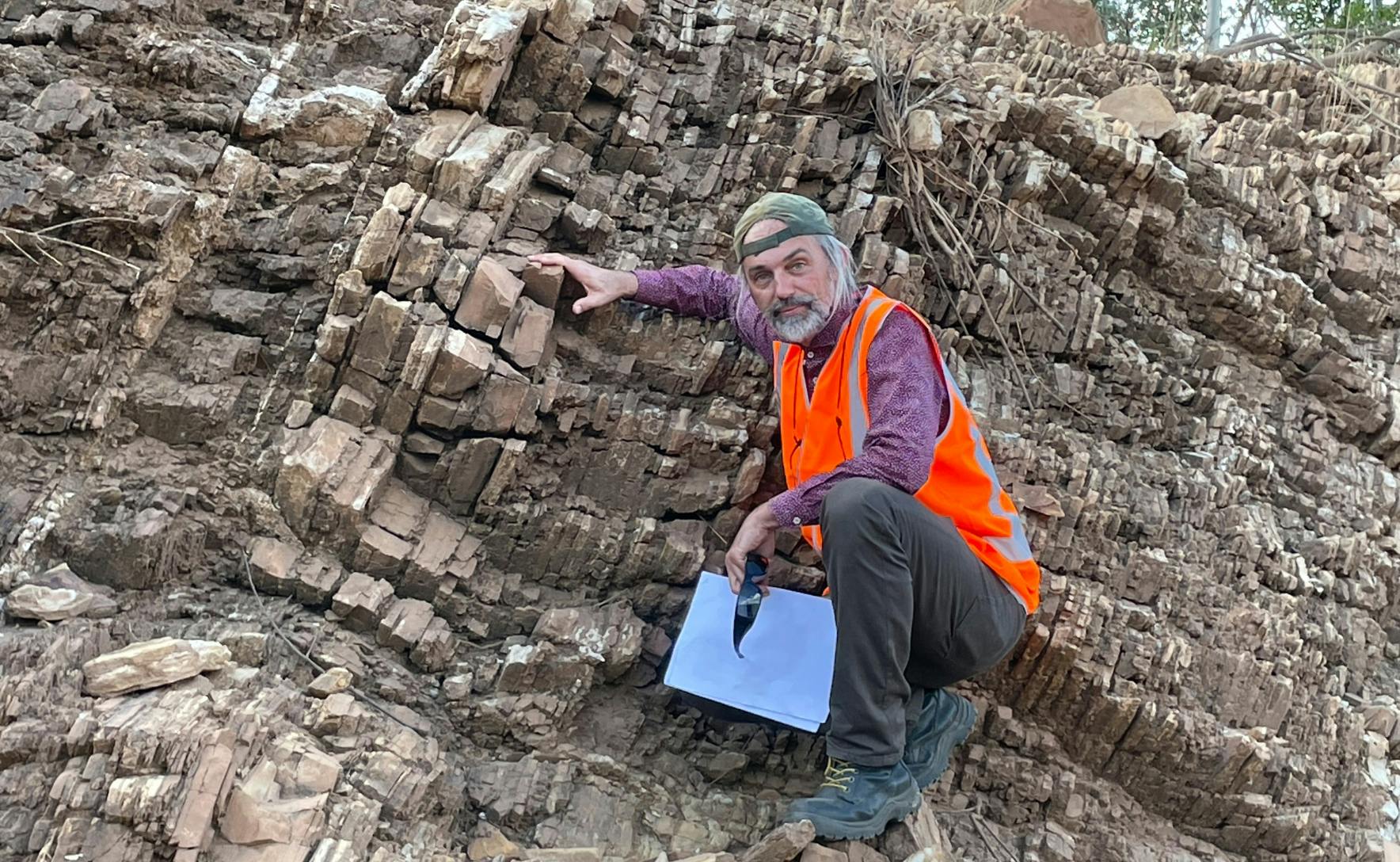 Professor Jochen Brocks inspecting the 1.6-billion-year-old rocks in Australia's Northern Territory. He is crouching next to the rock face and is wearing a fluorescent orange vest and a green backwards cap.