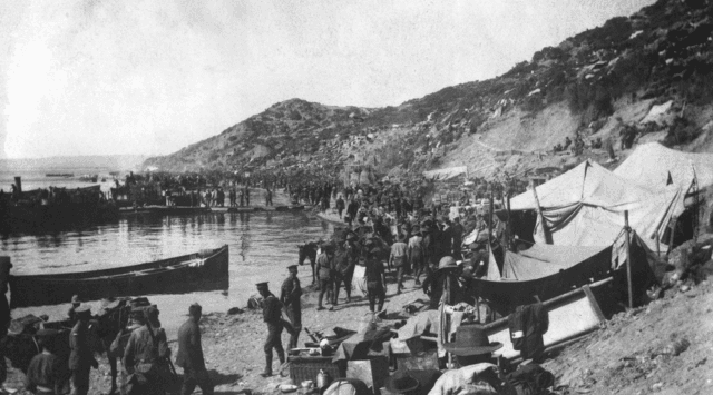 A grainy black and white image of soldiers on the shore of a beach. There are makeshift canvas tents on the sand and small wooden boats in the water.